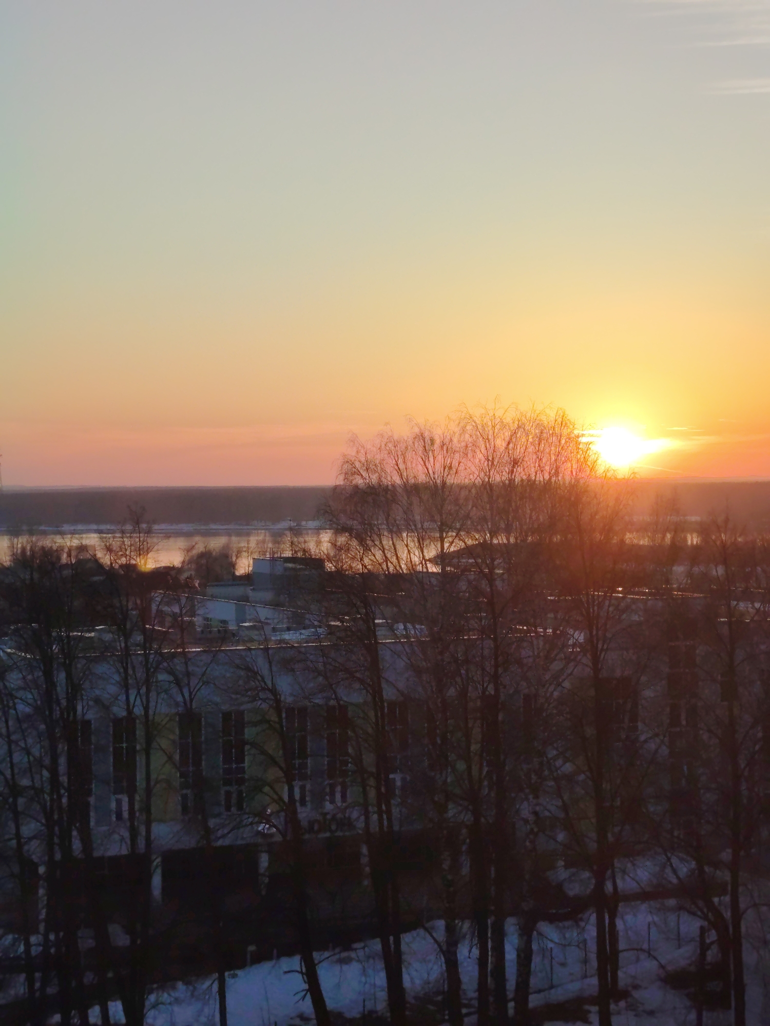 The image shows a cityscape at sunset, likely in the city of Chaikovsky, Russia. In the foreground are silhouetted bare trees partially obscuring a building with multiple windows. Beyond the building, there's a body of water or river with reflections of the setting sun. The sky is a gradient of colors, transitioning from a light, muted tone near the horizon to a brighter, more saturated orange where the sun is positioned. Patches of snow are visible on the ground. No people are visible in the image.