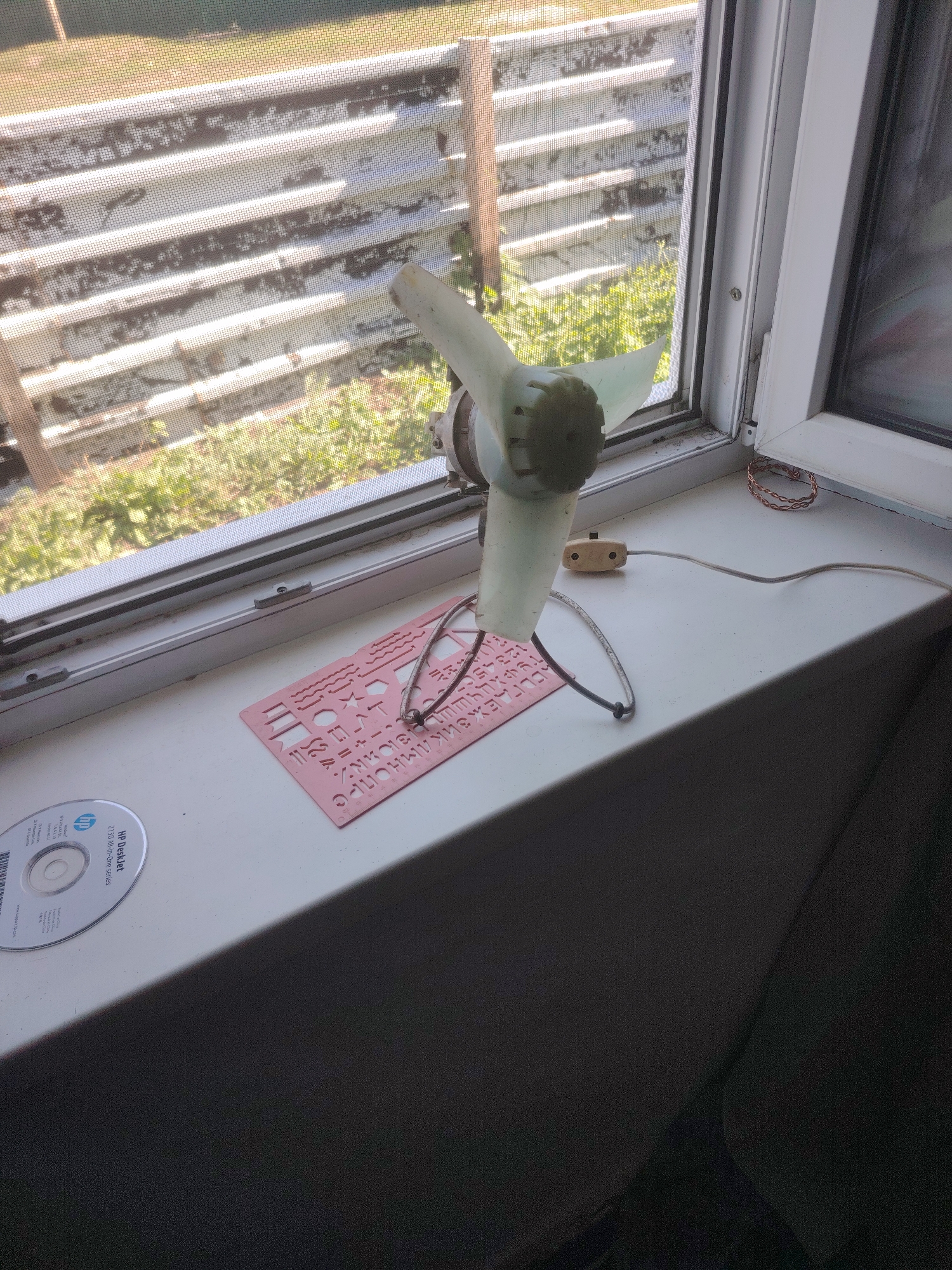 An old, cream-colored electric fan with bent blades is positioned on a white windowsill, facing towards the viewer's left. The fan's power cord is plugged into a wall outlet visible behind it. To the left of the fan, a pink plastic stencil with various geometric shapes and letters is laid flat. Further left, a silver CD with the HP Deskjet logo is visible. To the right of the fan, a coiled copper wire sits on the windowsill. The windowsill is located indoors, with a screened window behind the fan showing a view of what appears to be a rural outdoor setting. The window frame is white. The scene suggests an indoor space, possibly a room, overlooking an outdoor area in Александров ауылдық округі, Kazakhstan.