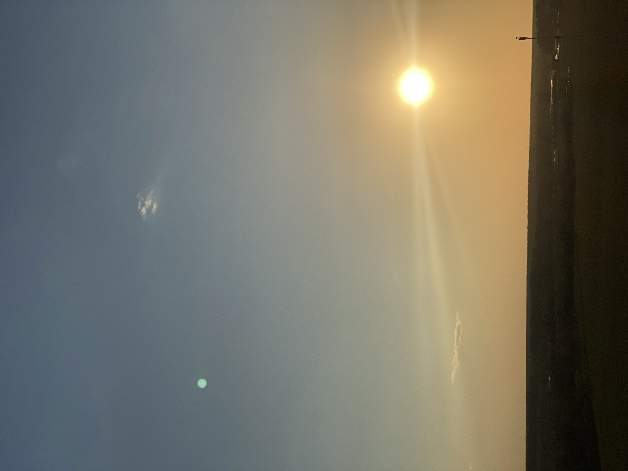 The image depicts a sunlit sky transitioning from blue to orange at the horizon. The sun is a bright, circular shape in the upper right portion of the frame. A thin line of land and trees is visible along the right edge. There are a few small clouds in the sky, with one small, bright green spot seemingly a lens flare. A lamppost is visible on the right edge. The location is Unknown, Unknown.