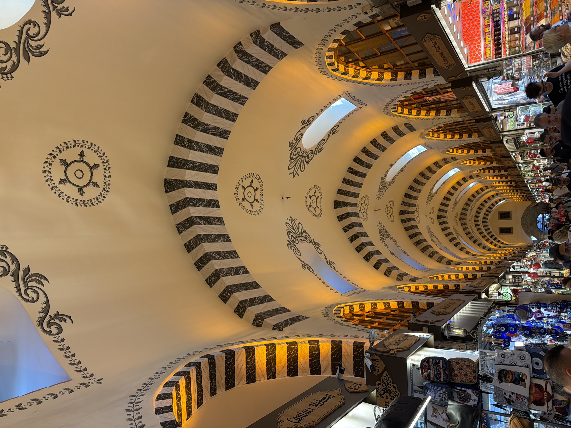 This is an indoor photograph taken from a low angle, looking up at the ornate ceiling of a marketplace. The ceiling is a prominent feature, with a large, curved surface adorned with decorative motifs. Bands of black and white chevron patterns alternate with plain white sections, creating a dynamic visual effect. Stylized floral and circular emblems are painted on the white surfaces, adding to the intricate design. Several skylights are visible along the curve of the ceiling, allowing natural light to filter in and casting a warm glow, suggesting it is daytime.
Below the ceiling, the scene opens up to reveal a bustling marketplace. Stalls are packed with various goods, including textiles, souvenirs, and decorative items like the blue evil eye charms seen in the foreground. Several people are browsing and shopping, their faces suggesting engagement and interest in the products. The overall atmosphere appears lively and vibrant. One stall has a wooden sign with the visible text "Contact Mehmet". The architecture and the goods on display suggest the location is likely a historic bazaar in Istanbul, Turkey. The image captures the essence of a traditional market experience, blending architectural beauty with commercial activity.