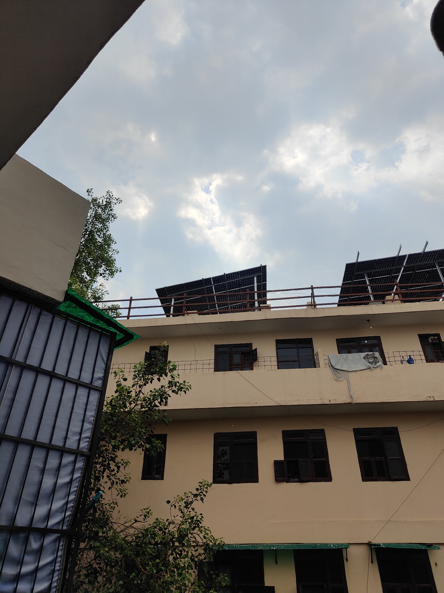 This image captures a building in Gurgaon, India, under a cloudy sky. The building features a light beige facade and has multiple floors. On the top floor, there is a railing and two sets of solar panels installed on the roof. The building has numerous windows, many of which appear to be dark-tinted or have dark frames. Some windows on the upper floors are open, while others are closed. A light-colored sheet is draped over a railing on one of the upper floors.
To the left of the building, a part of another structure is visible, with a gray exterior on the upper portion and a dark-colored grating or screen on the lower portion, covered with a green material. Trees with dense green foliage are growing near the building, with branches extending towards the structure and the sky. The overall scene suggests an urban or semi-urban environment in Gurgaon.