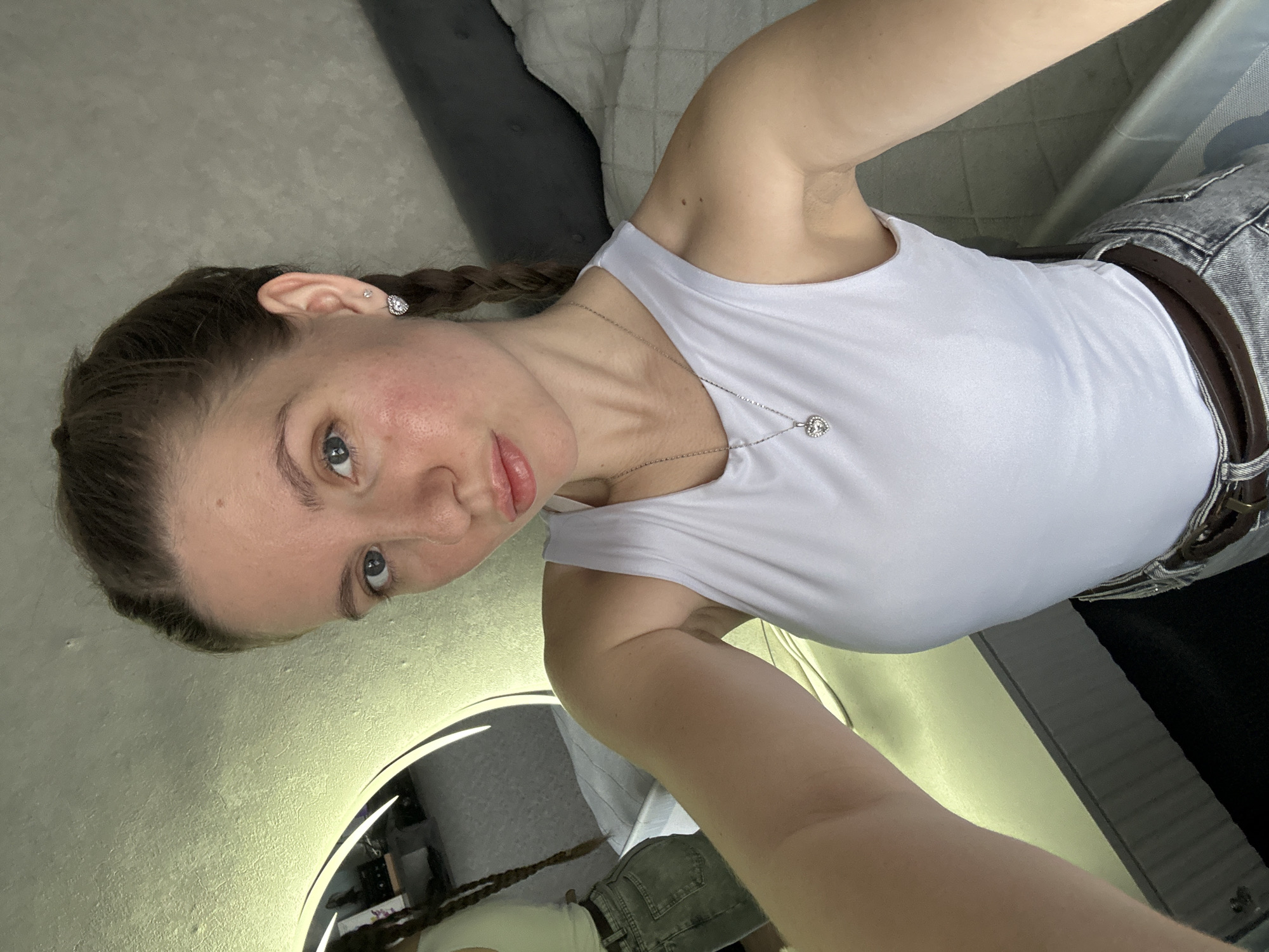 A young woman is shown in a selfie. She has light skin, blue eyes, and her hair is styled in braids. She is wearing a white tank top and a silver necklace. A brown belt is visible on her waist and she is wearing denim shorts. The background includes a round mirror with a bright light and parts of a room with a gray couch. It is not possible to verify the location is in the city of Klin, Russia.
