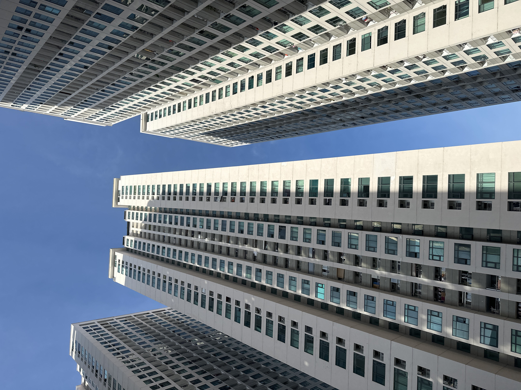 The image shows a low-angle view of several tall residential buildings against a clear blue sky. The buildings are modern in design, with light-colored facades and numerous windows with dark frames, many of which appear to be tinted or covered with light-colored curtains. Balconies are visible on some of the buildings, with some displaying laundry or other personal items. The buildings are positioned close together, creating a sense of density. The sunlight casts shadows on the facades, indicating the time of day. The environment appears to be urban. The location is identified as Bac Nha Trang Ward, Vietnam.