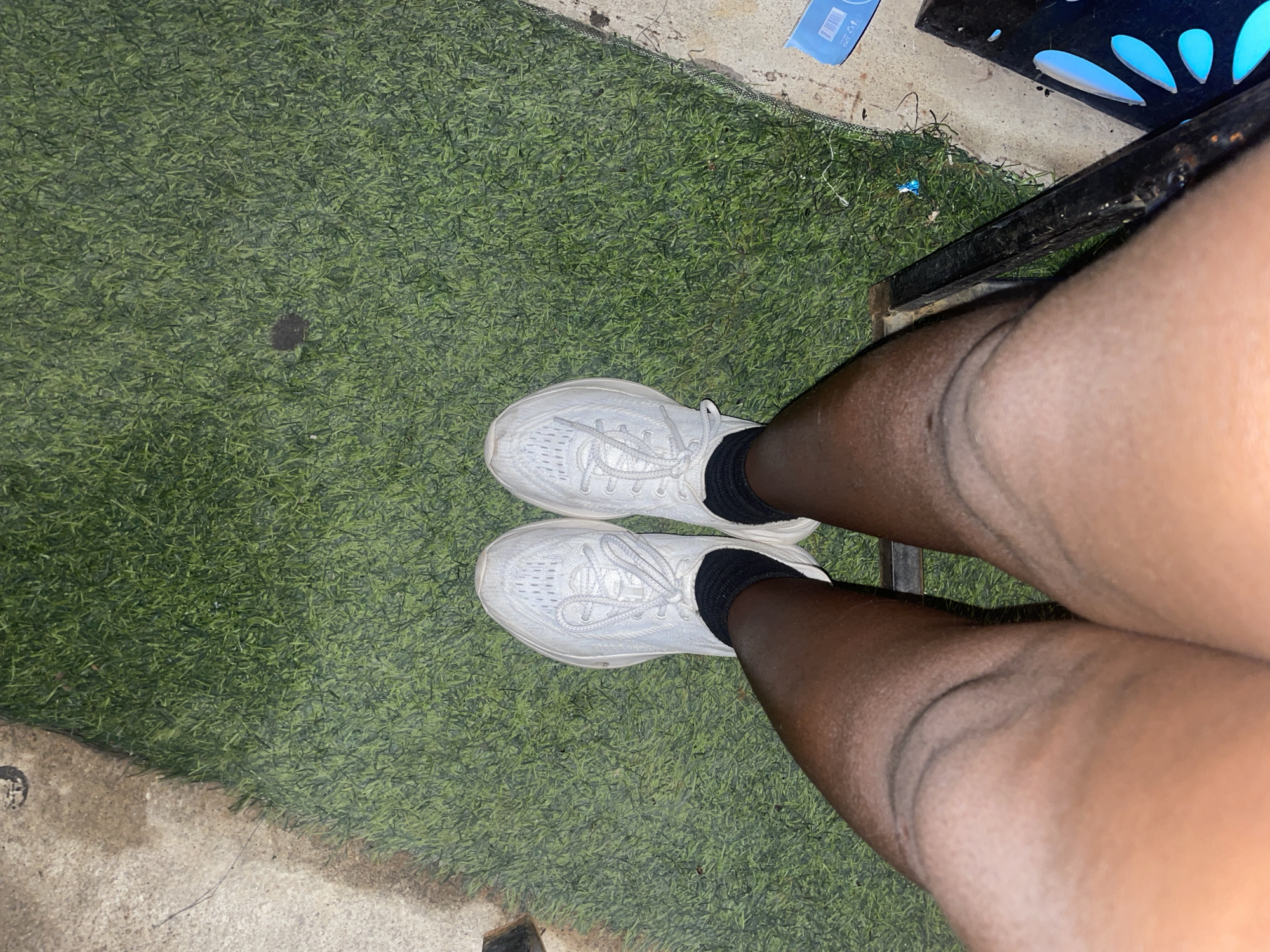The image shows a close-up, overhead view of a person's legs and feet, standing on what appears to be artificial turf. The person is wearing white athletic shoes and black socks. The skin tone is dark. The background consists of green artificial turf and some concrete surfaces. A black metal object is partially visible on the right side of the frame. There are no discernible city landmarks or identifying environmental features that would indicate a specific location within a city.