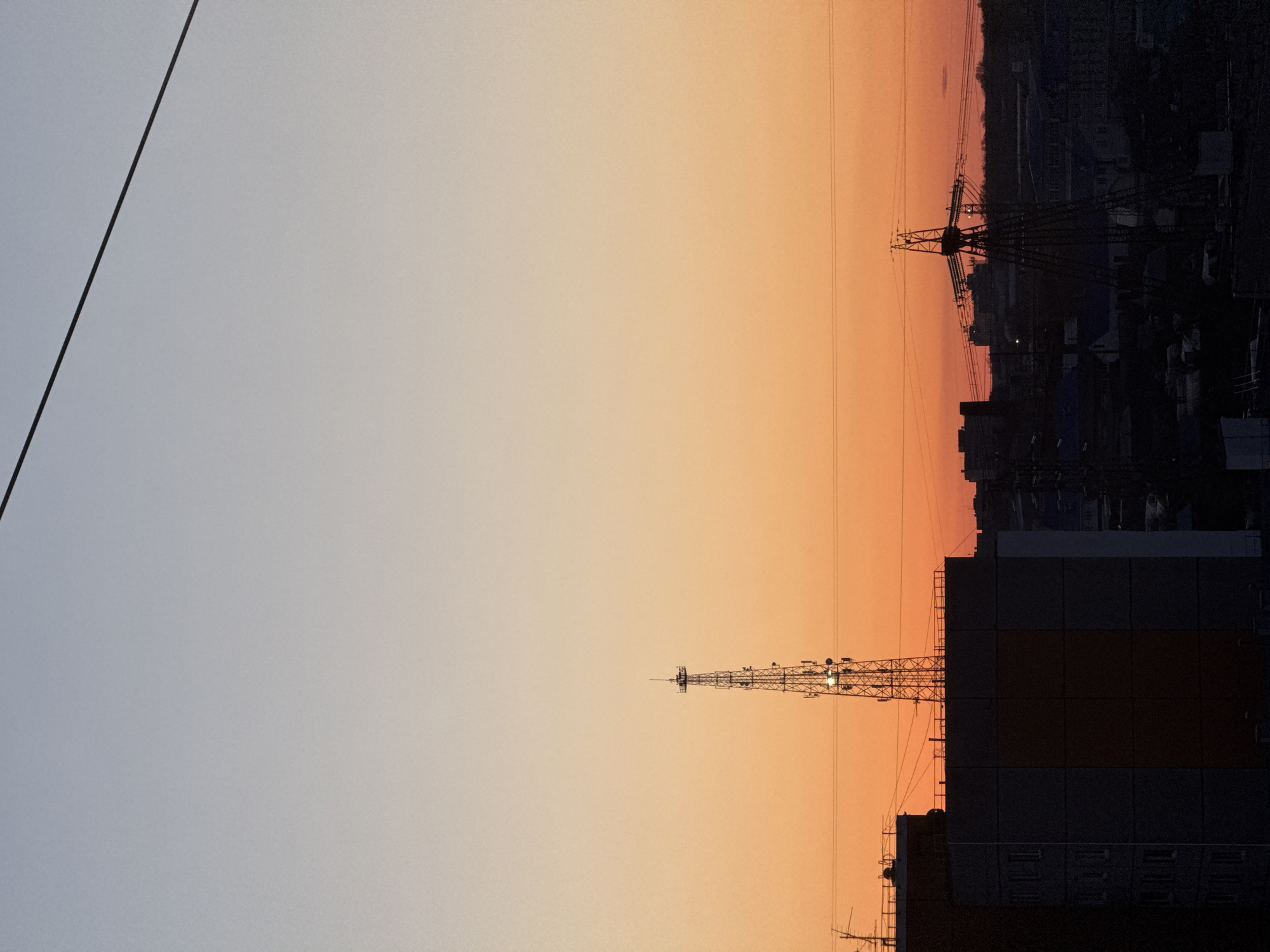 The image shows a cityscape at sunset, likely in Chaikovsky, Russia, as indicated by the presence of buildings and infrastructure typical of a city. The sky transitions from a light grey-blue on the left to a warm orange and red hue on the right, indicative of the setting sun. A black electrical wire cuts diagonally across the upper left portion of the frame. The right side of the image is dominated by the silhouette of buildings against the colorful sky. Prominent features include communication towers, power lines, and dense residential structures in the background. The overall lighting is dim, emphasizing the contrast between the sky and the darker foreground.