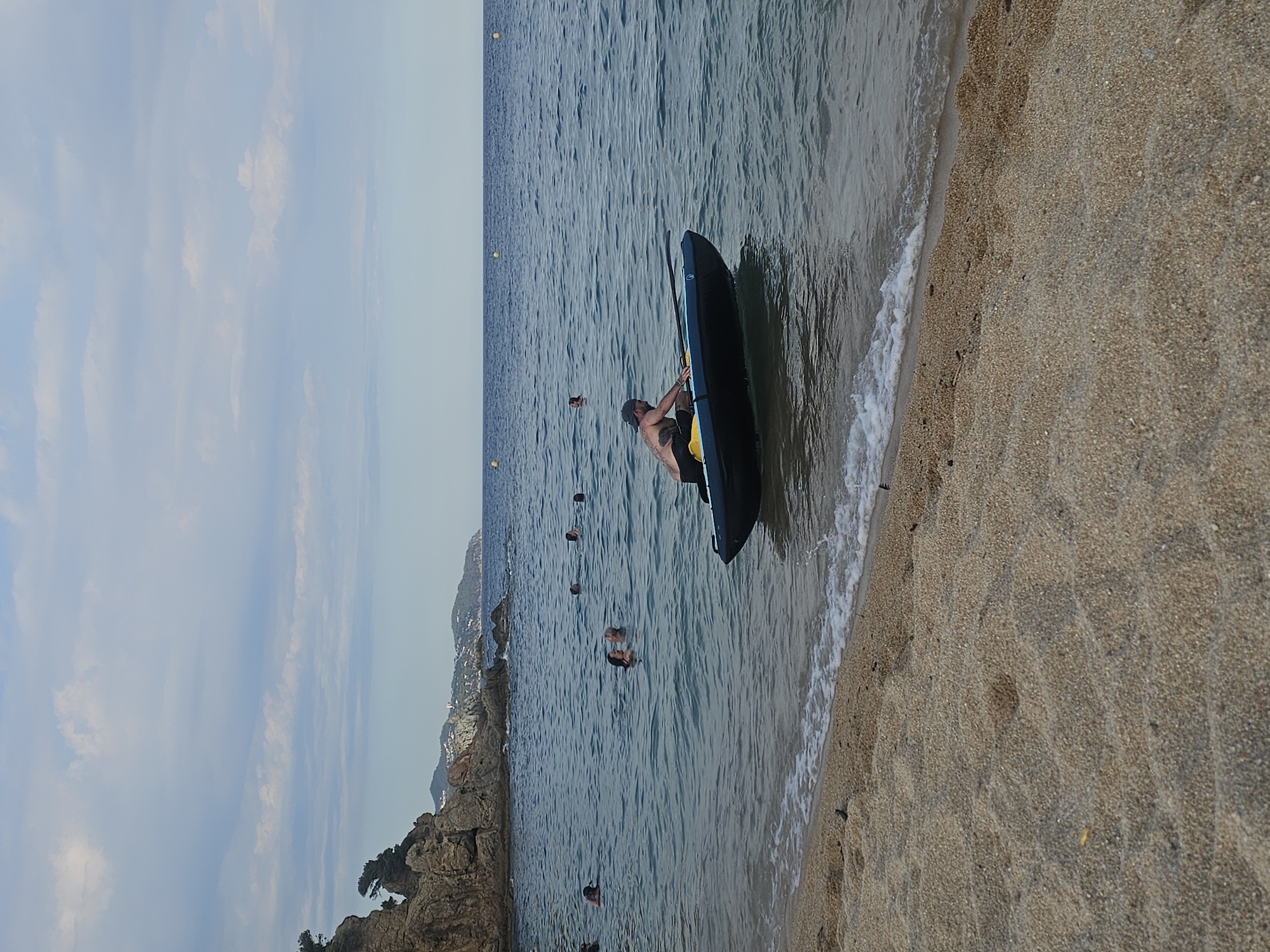 The image depicts a beach scene in Blanes, Spain. The primary subject is a person in a black kayak, positioned at the edge of the water where it meets the sand. The person is wearing a dark cap and yellow shorts, and appears to be preparing to enter or exit the kayak. The water is calm with gentle waves lapping at the shore. In the background, several other people are visible swimming or wading in the sea. Rocky outcrops and lush vegetation line the coastline in the distance, suggesting a scenic, natural environment. The sky is mostly clear with some wispy clouds, indicating fair weather, likely during the daytime. The overall mood is relaxed and recreational, as people enjoy a day at the beach. The sandy texture of the beach and the clear blue water are prominent details. No visible text is present in the image.