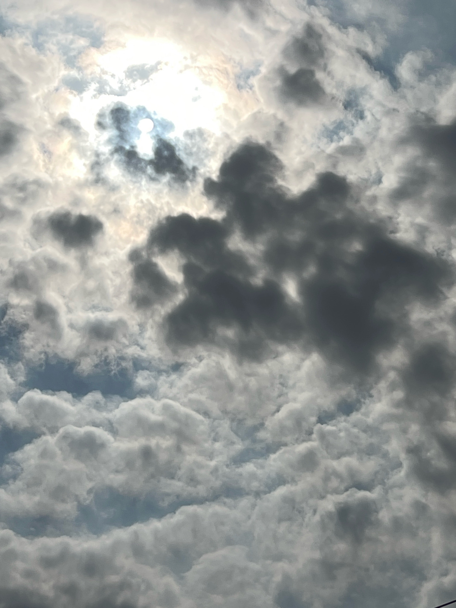 A close-up, low-angle shot captures a turbulent sky filled with dramatic, dark gray clouds partially obscuring a bright, hazy sun. The sun's disk is visible as a white circle through a break in the clouds, with a halo effect from the surrounding light. Wispy, lighter gray clouds mingle with the darker formations, creating a sense of depth and movement. Hints of blue sky are visible in the gaps between the clouds. The scene appears to be taken from ground level, looking directly upwards. There are no identifiable people, structures, or specific landmarks that would indicate the location as Kuta, Indonesia; the image exclusively depicts the sky and cloud formations.