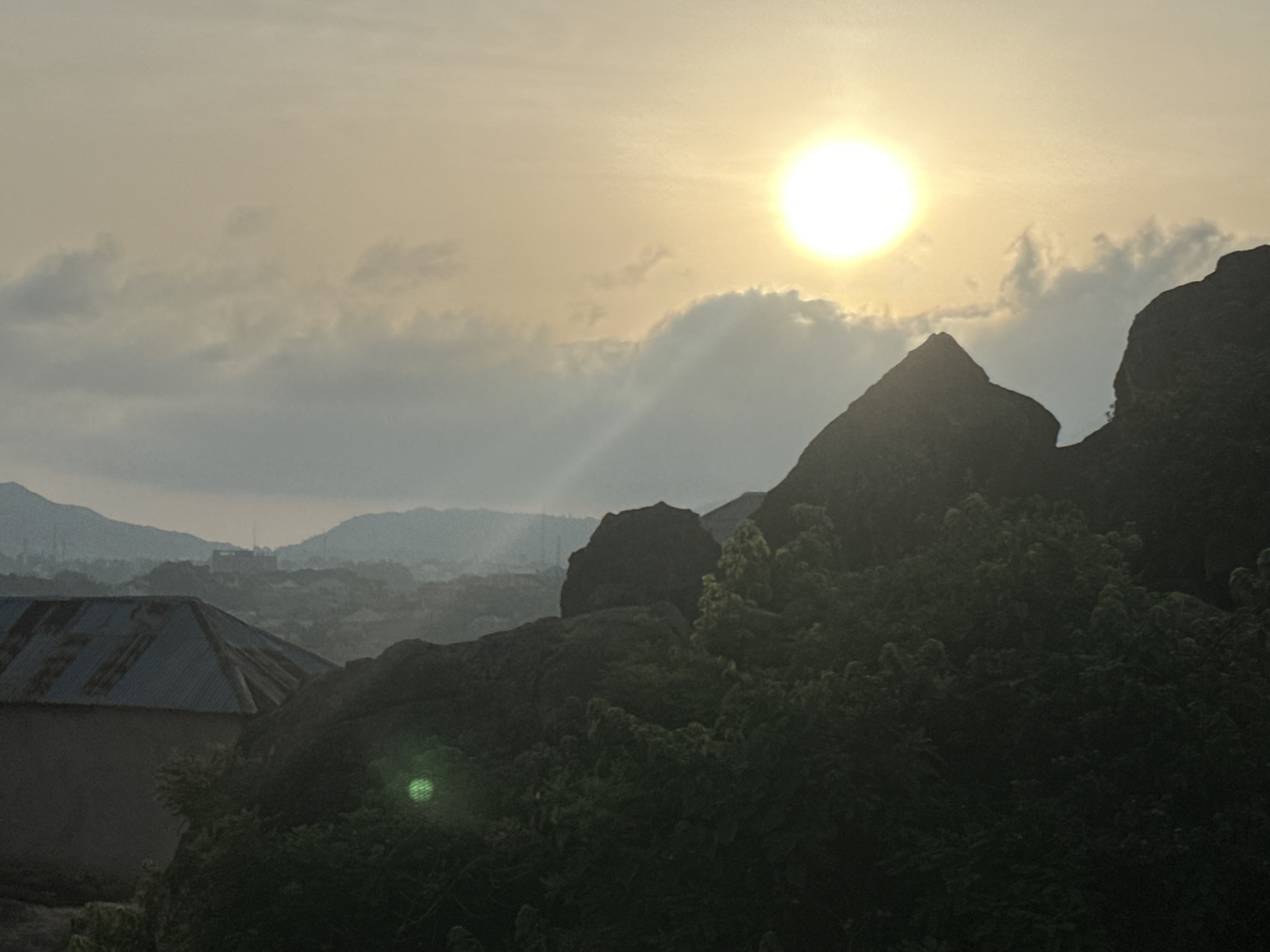 This wide-angle landscape image captures a scenic view in Jos, Nigeria, likely at sunrise or sunset. A brilliant, low-lying sun dominates the upper right of the frame, casting a warm, pale yellow-orange glow across the sky. Wispy clouds are scattered, with some partially obscuring the sun and creating subtle light rays.
In the mid-ground and foreground, rugged, dark rock formations and dense green foliage are silhouetted against the bright sky, emphasizing their dramatic shapes. A prominent building with a corrugated iron roof is visible in the lower-left corner. The backlighting creates a lens flare, seen as a green circular artifact in the lower-left among the trees.
The background reveals a hazy vista of rolling hills and distant mountains, with faint outlines of a town or city nestled in the valley, suggesting human settlement within the natural terrain. The atmospheric perspective contributes to the sense of depth. No people or visible text are present. The image evokes a serene and atmospheric mood, highlighting the distinctive rocky landscape of the Jos Plateau during a golden hour.