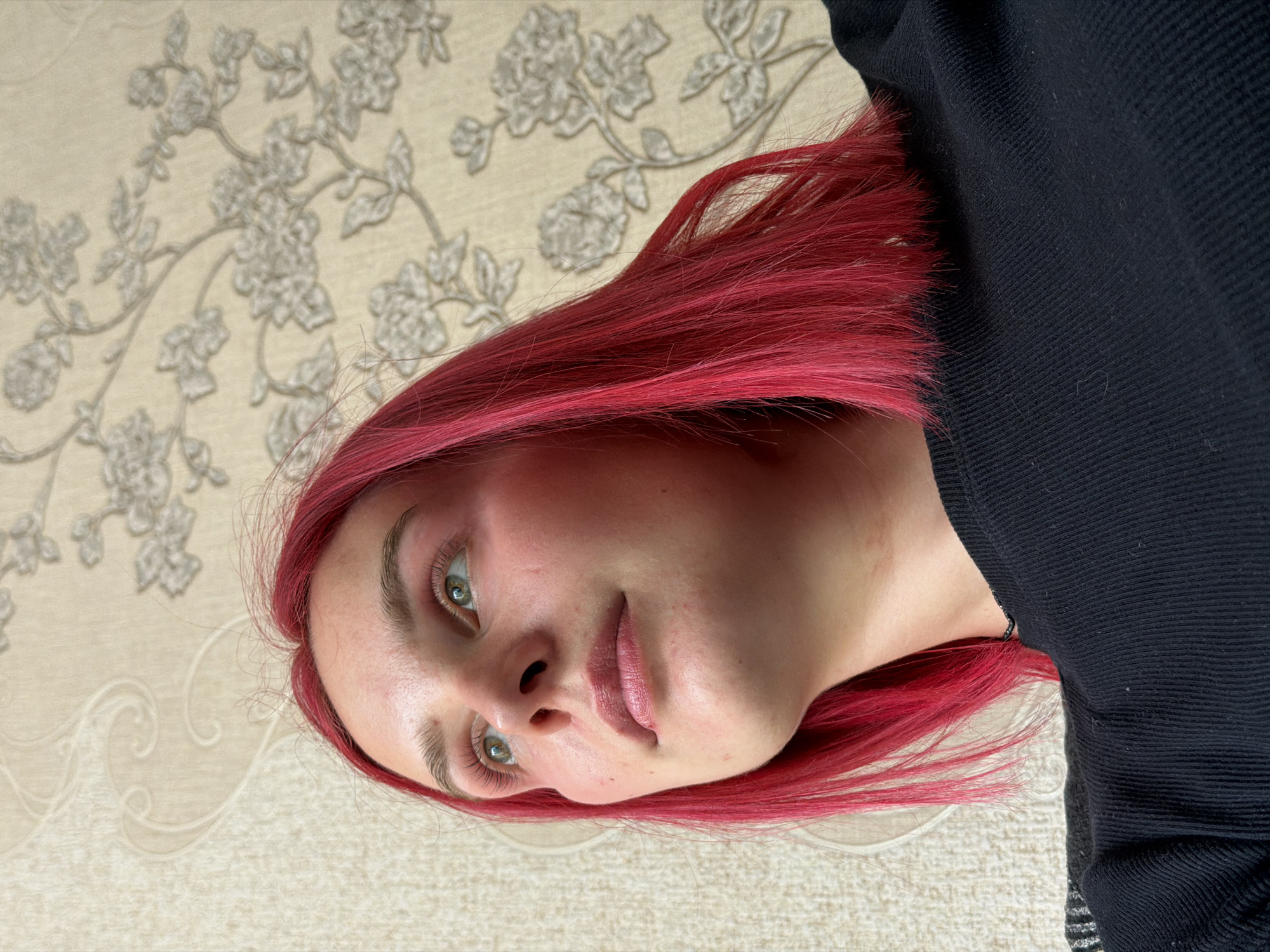 A close-up portrait features a young woman with striking, long, bright red hair. She is positioned with her head tilted upwards and slightly to the right, looking towards the viewer with her green eyes. Her expression is calm and neutral, with her lips slightly parted. Her skin appears fair, with subtle redness on her cheeks, and she has visible eyelashes.
She is wearing a dark, possibly black, long-sleeved garment with a ribbed or textured pattern, visible on her right side, extending from her shoulder area. The background behind her is a light, cream-colored surface, possibly wallpaper or fabric, featuring an embossed or patterned design of delicate floral branches and leaves in a lighter shade, creating a subtle contrast. Another section of the background shows a less distinct wavy pattern at the bottom. The lighting suggests daytime, casting soft shadows that highlight the textures of her hair and the background. There is no visible text in the image. The overall scene conveys a casual, intimate portrait.