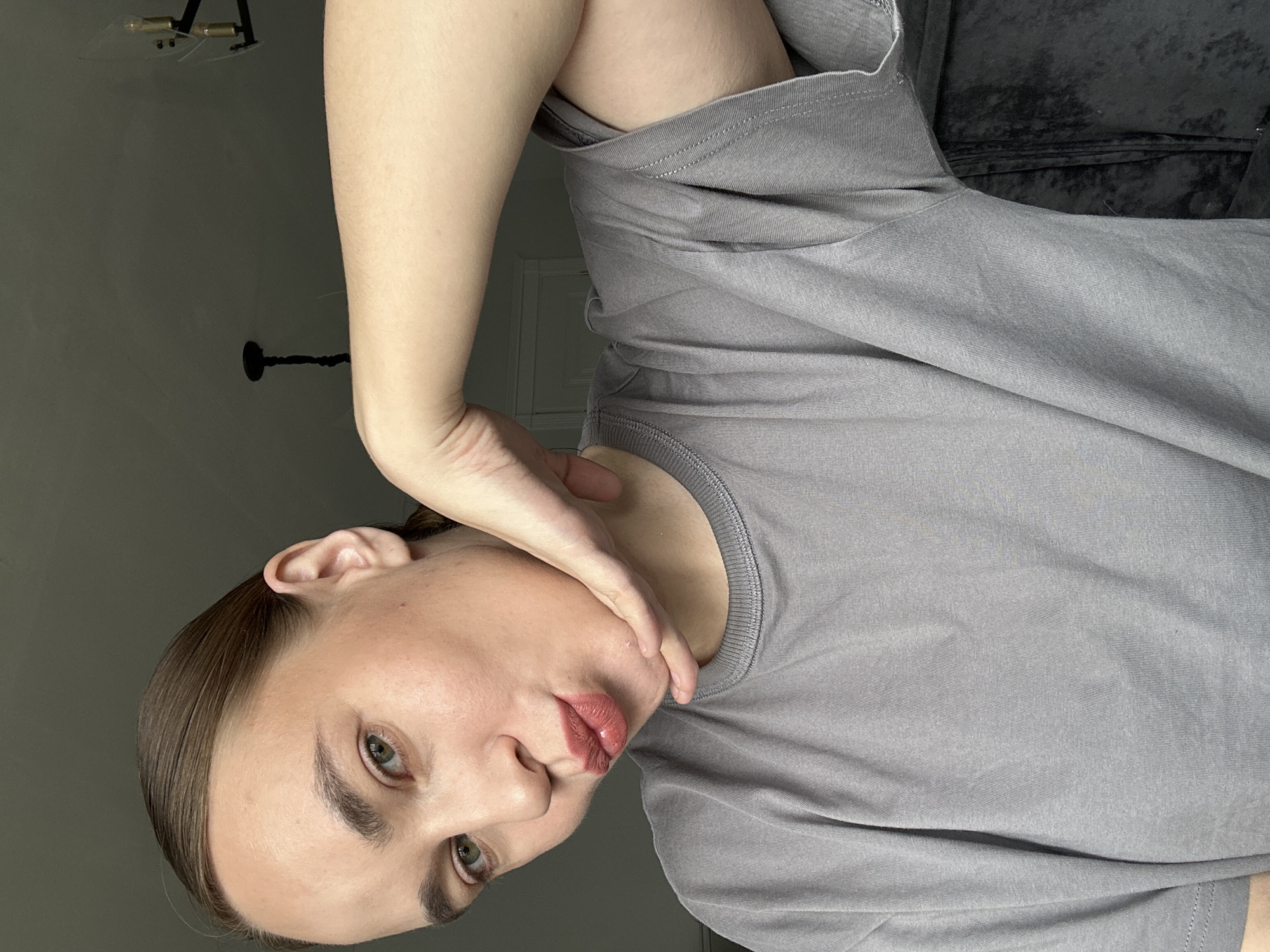 A woman is posing for a portrait. She is fair-skinned with brown hair pulled back. Her eyes are green and her lips are red. She is wearing a grey t-shirt. Her left arm is raised with her hand touching her neck. The background is a gray wall. A portion of a chandelier is visible in the top left corner. The location, Kharnavy, Russia, is not supported by visual evidence.