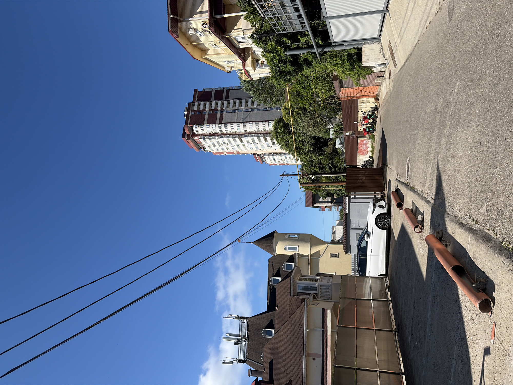 The image depicts a residential area in Sochi, Russia, on a clear, sunny day. In the foreground, a street runs from the bottom right towards the middle distance. The street is paved and appears to be a quiet residential road. Orange pipes, possibly for construction or drainage, are lying on the side of the road. To the left of the street, there is a paved area that might be a parking lot, with a white car parked there. Beyond the parking area, there are several buildings. One building has a distinctive conical tower and a dark brown, pitched roof topped with multiple antennas. Another building is a tall, multi-story residential building with a facade of white and maroon panels. To the right of the street, there are more residential buildings with yellow siding and balconies, surrounded by lush green foliage. In the background, the clear blue sky is visible, with a few wispy clouds. The overall impression is of a sunny, warm day in a coastal city.