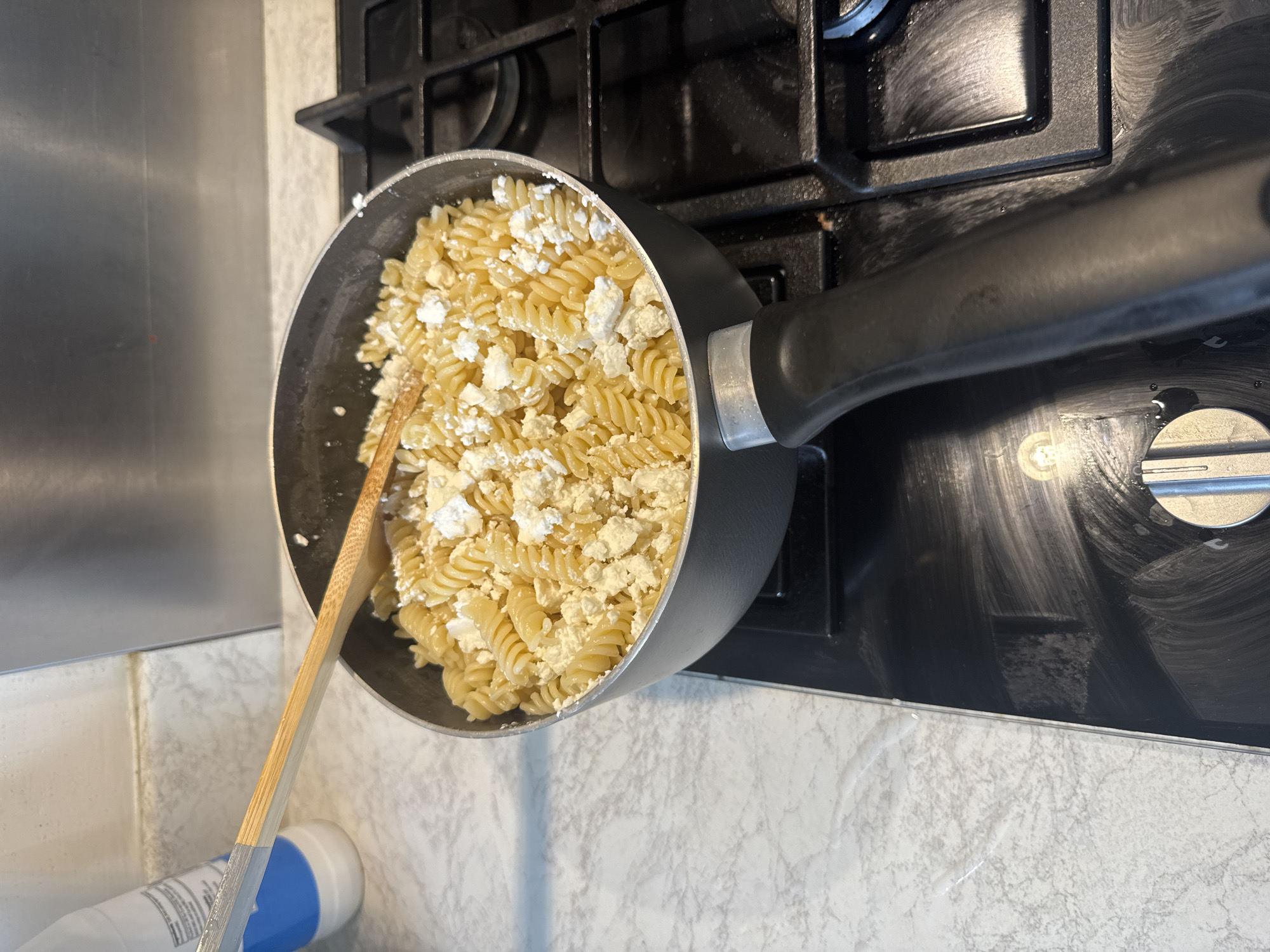 A top-down shot shows a black saucepan filled with cooked fusilli pasta and chunks of white cheese. A wooden spatula rests inside the pan. The saucepan sits on a black glass stovetop. The stovetop is situated on a countertop featuring a white marble-like pattern. A portion of a white plastic bottle with a blue cap is visible in the lower left corner. The setting is likely a domestic kitchen in Swindon, United Kingdom. The focus is on the food preparation. No people are visible.