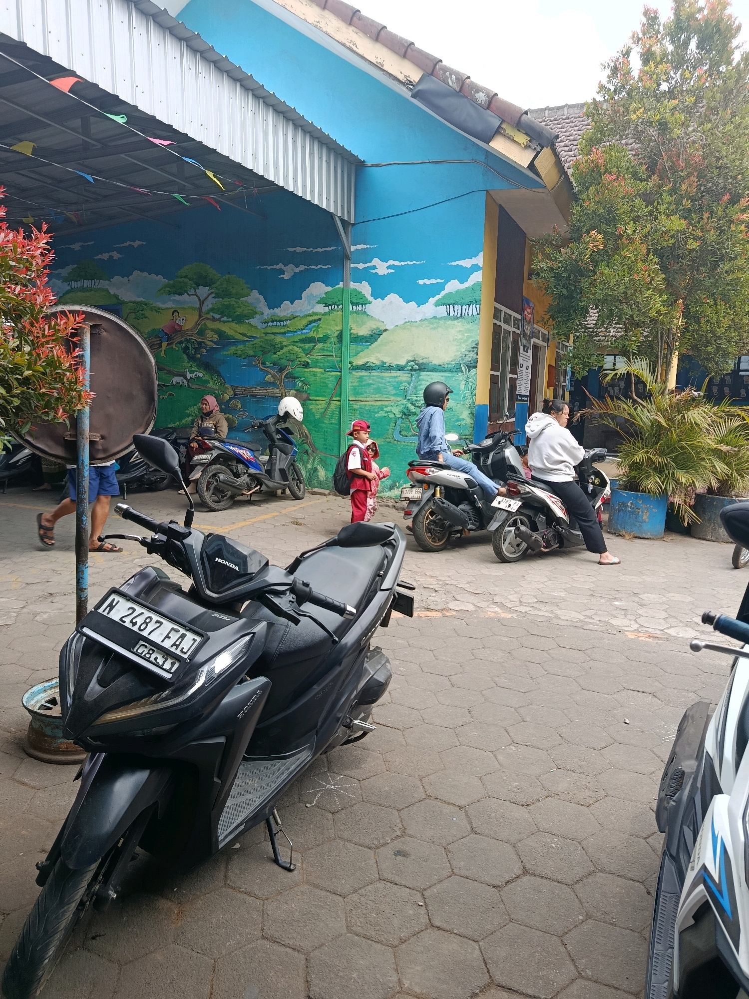 In front of a brightly painted building with a mural depicting a serene landscape, several motorbikes are parked on a hexagonal-tiled pavement. A black Honda motorbike with the license plate "N 2487 FAJ" is prominently displayed in the foreground. In the background, people are seen interacting near their parked motorcycles. A woman in a hijab is sitting on a motorbike, while another person in a blue denim shirt and jeans is seated on a different motorbike, with their leg casually extended. A child in red and white attire, wearing a backpack, stands near a motorbike. The building itself features a blue facade adorned with a vibrant mural and a yellow accent on its side. Lush green trees and foliage are visible in the background, contributing to the outdoor setting of this scene, likely in Bululawang, Indonesia.