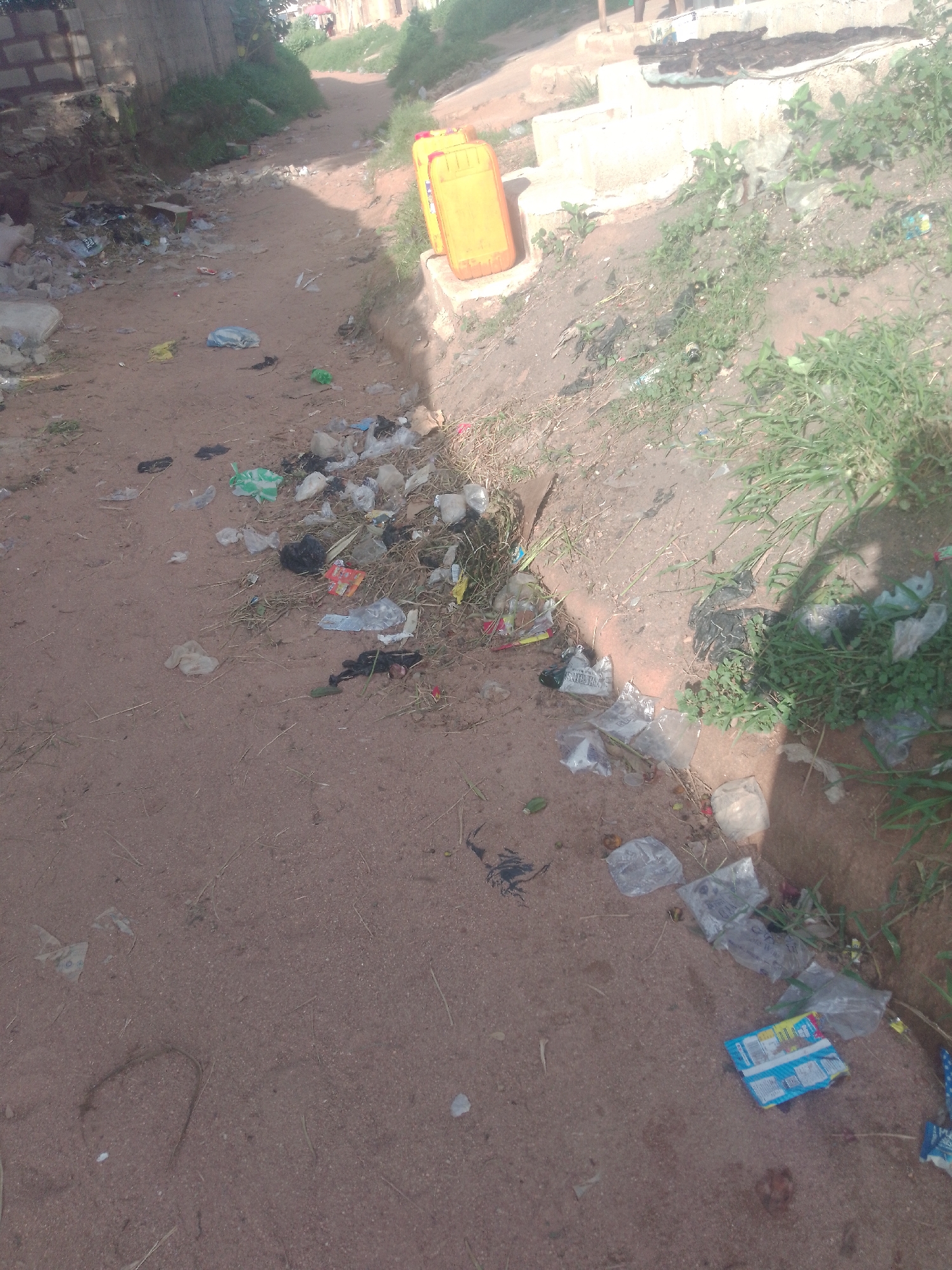 The image depicts a dirt path in Jalingo, Nigeria, heavily littered with refuse. The path is unpaved and uneven, with areas of dry grass and soil. On the left side of the path, a brick wall forms a boundary, with some vegetation growing alongside it. Further along the path, two large yellow jerrycans are visible, positioned on a raised concrete structure. The ground is strewn with a variety of waste, including plastic bags, wrappers, bottles, and what appears to be food remnants. Some of the plastic is translucent, while other items are opaque and colorful. The overall impression is one of neglect and unsanitary conditions, suggesting a public space that is not well-maintained. The light suggests it is daytime, and the shadow of a person or object is cast on the right side of the image.