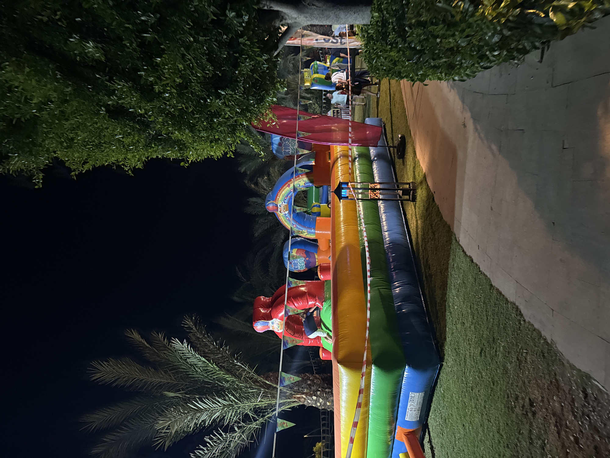A nighttime outdoor scene depicts a large, multi-colored inflatable playground with various structures, including a red climbing tower and a rainbow archway. A young child is visible on the red structure, reaching out. The inflatable is surrounded by green artificial turf, with a paved walkway running alongside. In the background, palm trees and lush green foliage are illuminated by artificial lighting. Several people are standing or walking around the perimeter of the playground. Decorative banners are strung across the scene. The overall atmosphere suggests a public event or entertainment venue. The location is identified as Aksu, Turkey.