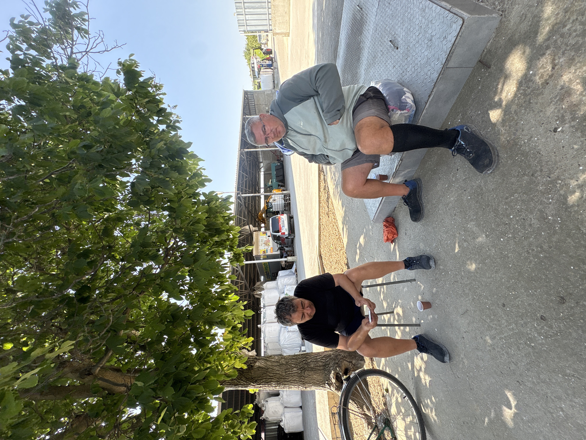 Two men are sitting outdoors near large white sacks under a tree. One man is on the left, sitting on a stool, wearing a black t-shirt and shorts, and looking at his phone. A bicycle is partially visible to his left. A small paper cup is on the ground in front of him. The other man is on the right, sitting on a metallic platform, wearing a grey and green hooded jacket, grey shorts, and black compression socks. He is looking towards the camera. In the background, there are several industrial buildings, some vehicles, and more white sacks. The ground is paved and appears to be an outdoor industrial or agricultural area. The sky is clear and blue. The location is identified as Iskra, Bulgaria.