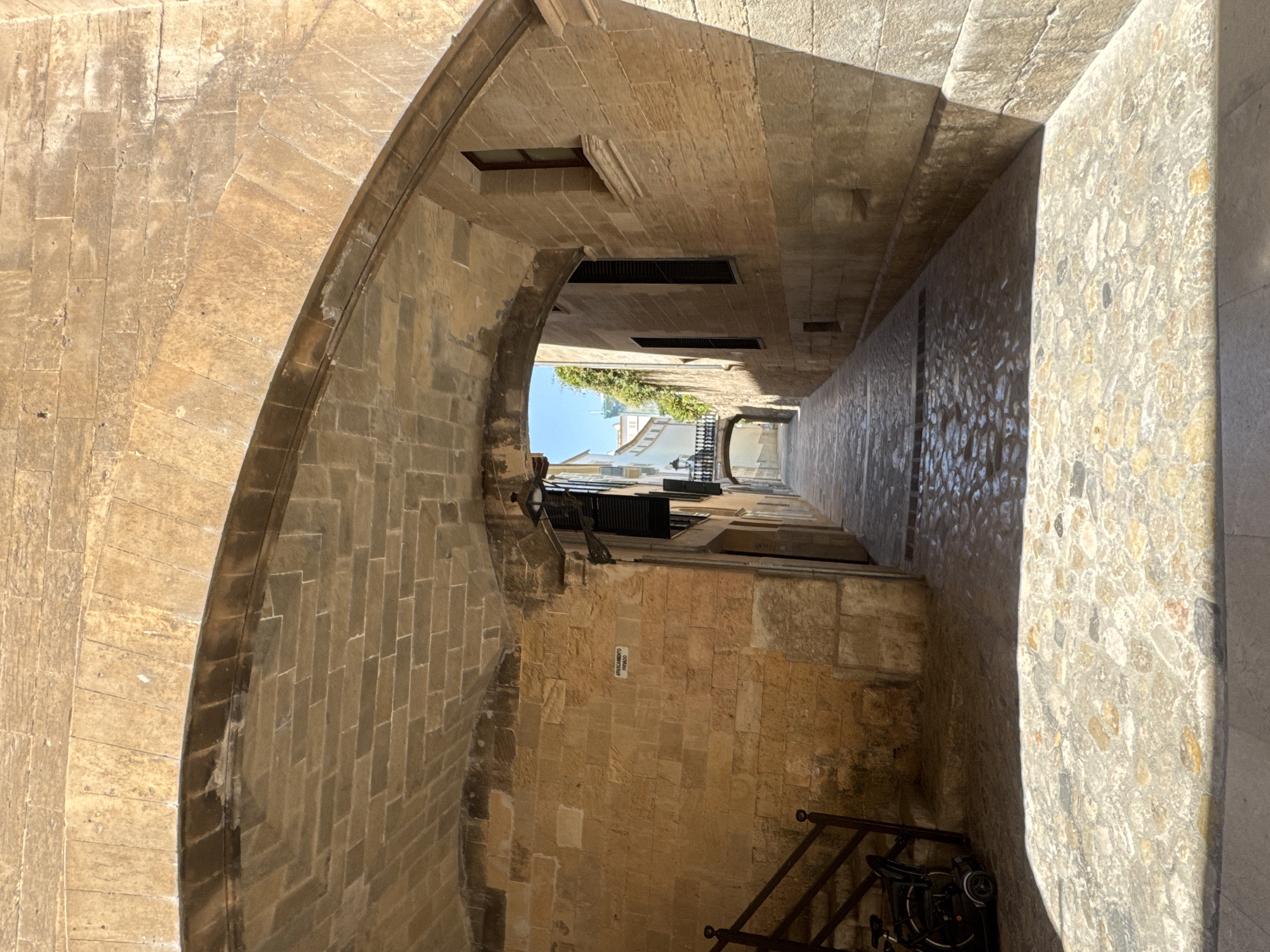 The image shows a stone archway in Palma de Mallorca, Spain, leading into a narrow cobblestone street. The architecture is historic, with rough-hewn stone walls and a vaulted ceiling in the archway. Sunlight casts strong shadows on the street, highlighting the textures of the cobblestones. To the left, a metal railing leads up a few steps, and a folded mobility scooter is parked at the bottom. On the wall to the left, a small white sign reads "APARCAMIENTO PRIVADO," indicating a private parking area. The street recedes into the distance, with buildings visible on either side. In the background, another stone archway can be seen, framing a bright sky and some greenery. The overall scene conveys a sense of timelessness and the charm of old European cityscapes.