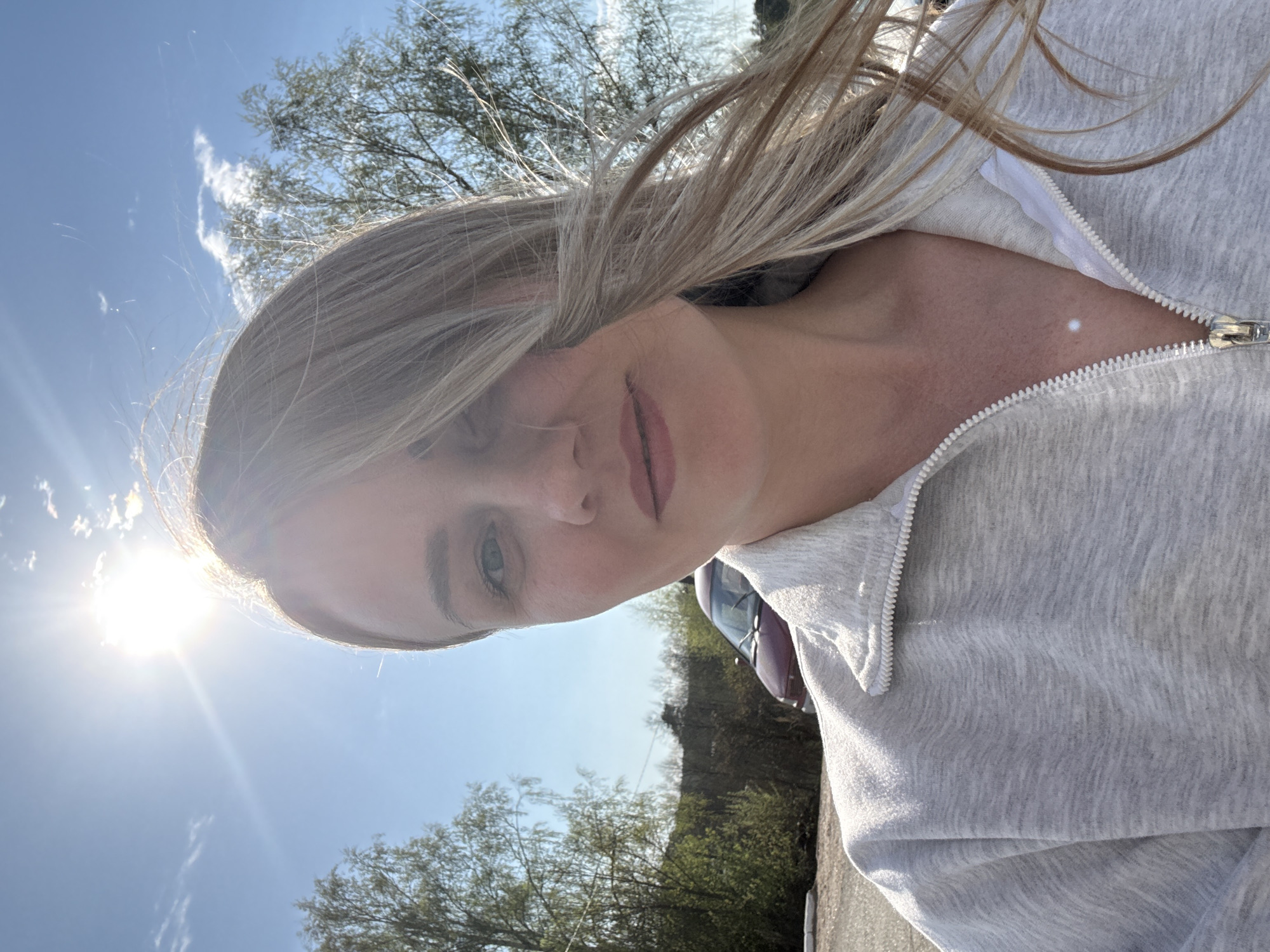 A woman with blonde hair, wearing a gray zippered hoodie, is outdoors on a sunny day. Trees are visible in the background, and the sky is blue with some clouds. The sun is bright in the lower left corner of the frame. A car is partially visible in the background. The location cannot be determined to be in Chaikovsky, Russia based on visual evidence.
