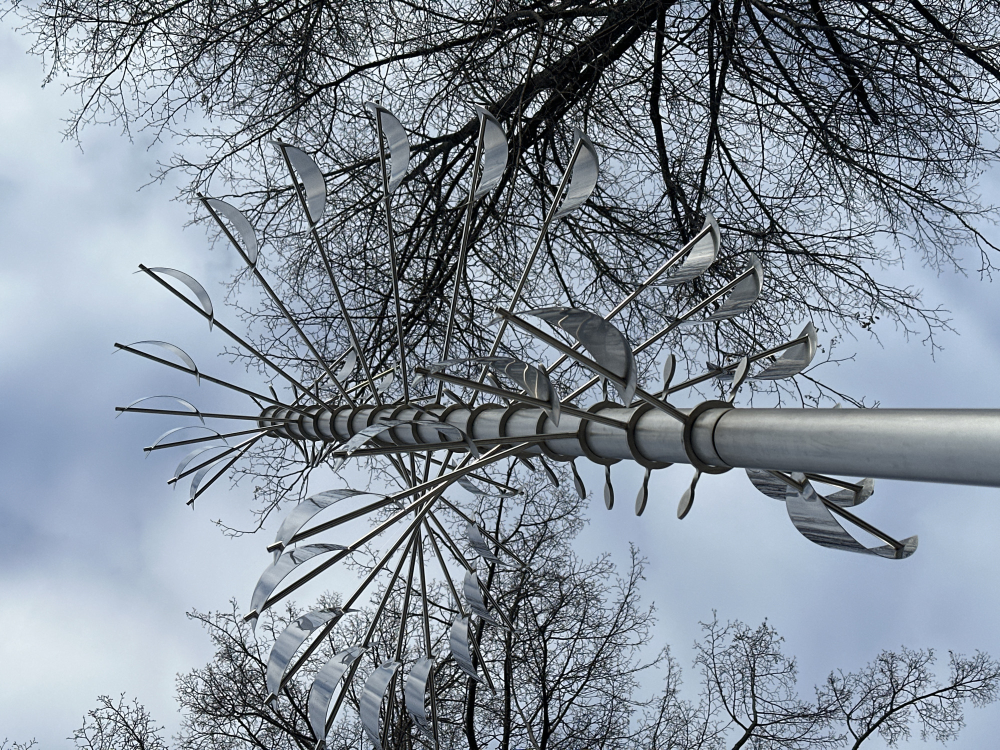 The image shows a kinetic sculpture in Votkinsk, Russia, viewed from a low angle looking up. The sculpture is made of silver metal and consists of a central, horizontal cylindrical pole with a series of rings. Thin metal rods extend outwards from the pole, each ending in a curved, crescent-shaped metal piece, creating a windmill-like effect. The sculpture is set against a backdrop of bare tree branches and an overcast sky. The branches of the trees fill the top and bottom portions of the image, framing the sculpture. The sky is grey with hints of blue.