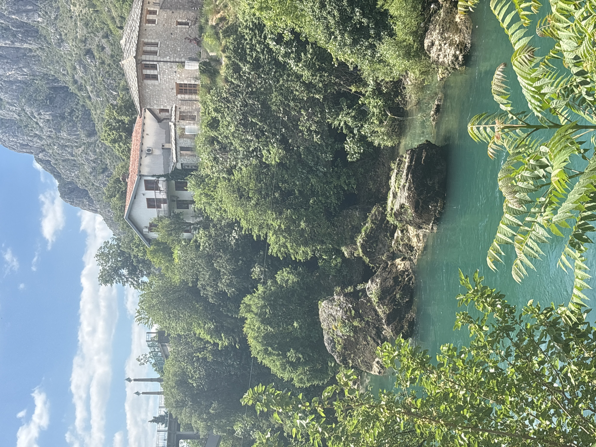 The image shows a view of the Buna River in Blagaj, near the city of Mostar, Bosnia and Herzegovina. The river has a distinctive blue-green hue. On the left side of the frame, a stone building, possibly a historic house or Tekke (monastery), is nestled between a rock face and dense green trees. In the foreground, foliage obscures parts of the view, adding depth to the composition. A section of a stone bridge or elevated platform is visible on the lower left side of the frame. The sky in the distance is blue with scattered clouds.