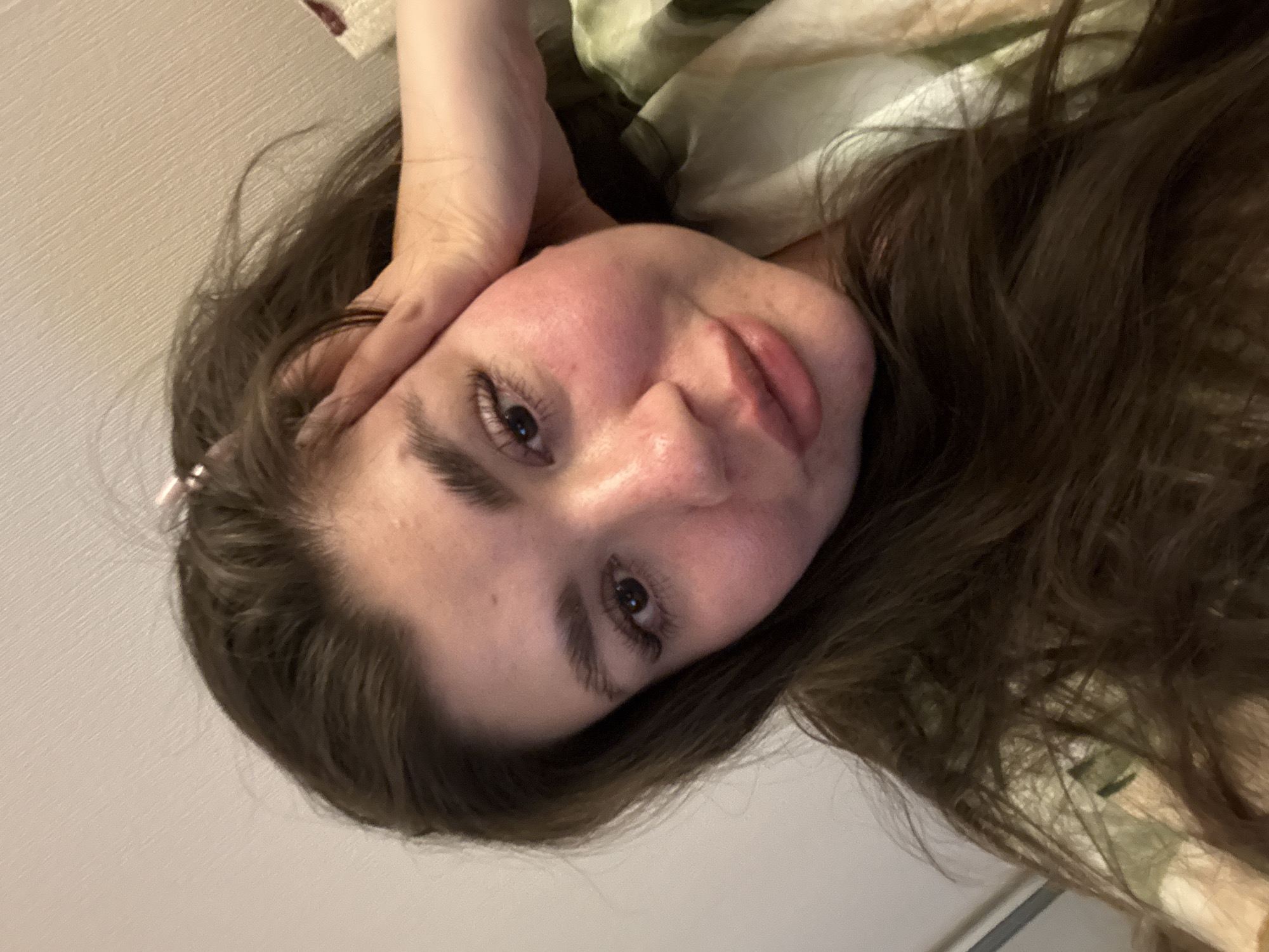 A close-up, angled selfie of a fair-skinned individual with brown hair, resting their head on their hand. The background includes a plain white wall and a floral-patterned blanket or sheet. No information is present in the image to suggest the location is in the city of Votkinsk, Russia.