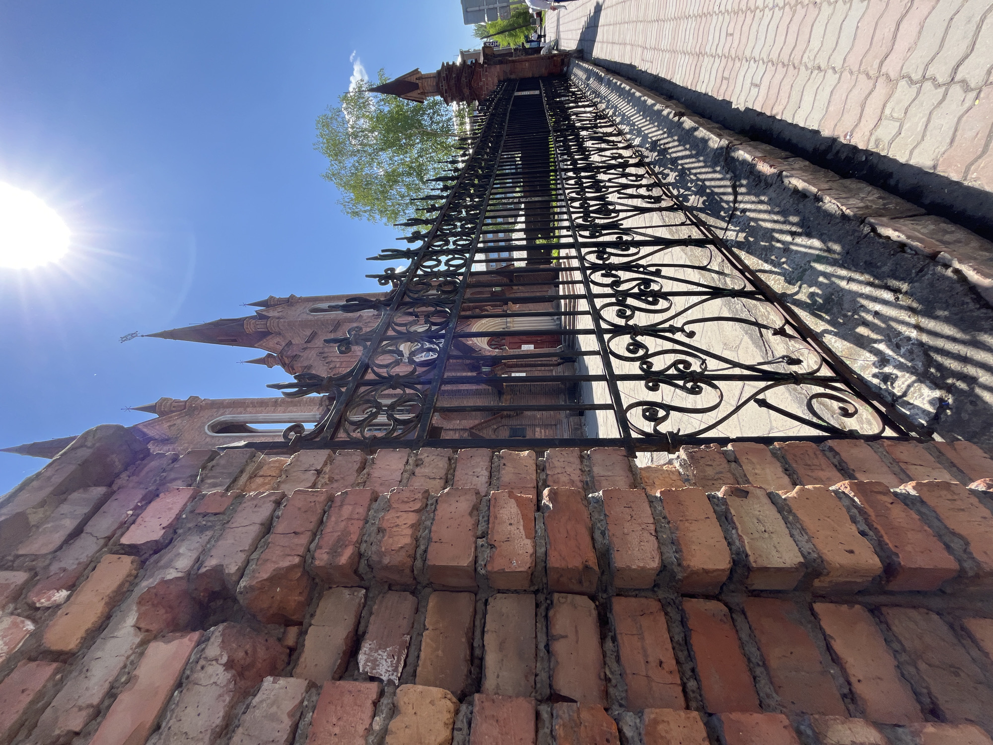 The image presents a low-angle, distorted view of a brick wall and an ornate metal fence, likely situated near a building in Krasnoyarsk, Russia. The lower portion of the frame is dominated by a rough brick wall, constructed of weathered red bricks with visible mortar. Above the wall, a black, wrought-iron fence extends across the scene. The fence exhibits decorative curls and pointed finials. Behind the fence is the building, potentially a church or historical site, identifiable by its red brick architecture and spire-like structures. A tree with green foliage is partially visible behind the building. The upper part of the image reveals a bright blue sky, with the sun glaring on the left side. A paved walkway, consisting of interlocking stone pavers, extends into the distance on the right. The perspective is skewed, creating a sense of depth and emphasizing the height of the wall and fence.