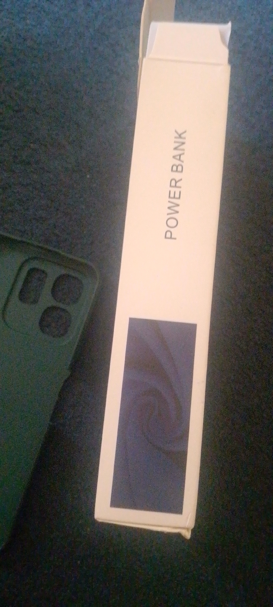 A power bank box with the words "POWER BANK" printed vertically on it is placed on a dark surface next to a dark green phone case. The box is open at the top. The phone case has a cutout for a camera. The surface appears to be carpeted.