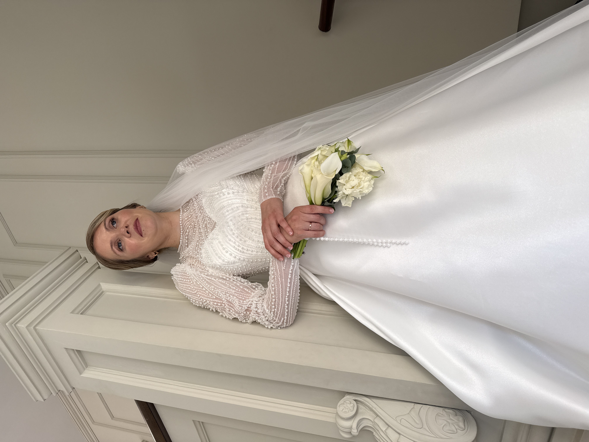 A Caucasian woman wearing a white wedding dress and veil stands with her back against a white staircase, holding a bouquet of white flowers. The dress is adorned with pearls and intricate beading on the sleeves and bodice. The woman's hands are clasped, with a ring visible on her left ring finger. She looks upwards and to the right, with a thoughtful expression. The background consists of the staircase and a neutral-colored wall. The location is not specified as Izhevsk, Russia.