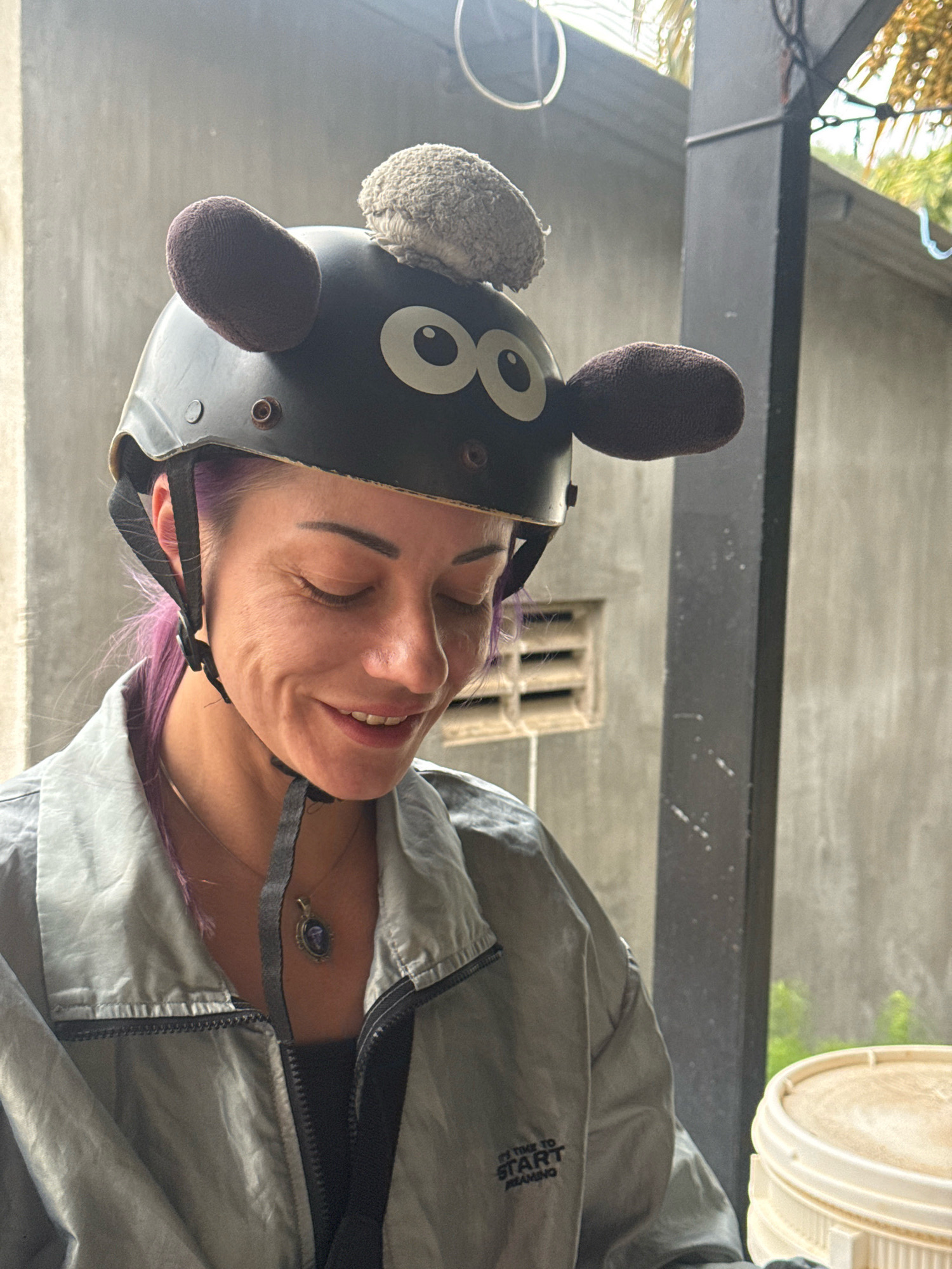 A woman with purple hair and a black helmet decorated with sheep ears and a fluffy head is looking down and smiling. She is wearing a gray jacket over a dark top and a necklace with a pendant. The background is a gray concrete wall with a vent and some greenery visible through a doorway. The scene appears to be outdoors, possibly in a covered area.