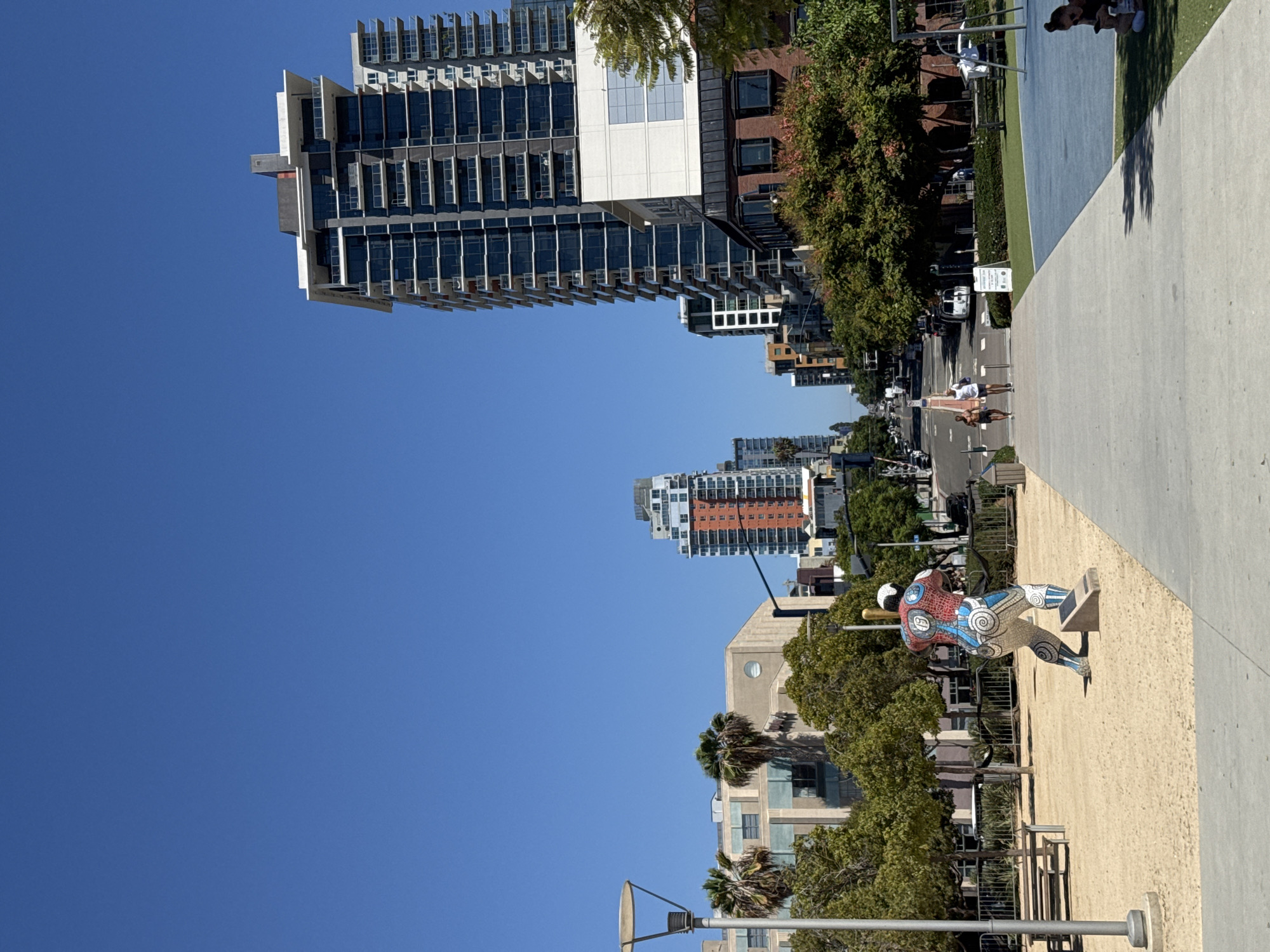 The image captures a street view in the city of San Diego, California, United States of America. In the foreground, a mosaic sculpture of a baseball player in mid-swing is prominently displayed on a sandy area. To the right, a paved path leads towards a park area with blue and green surfacing. In the midground, two individuals are running on the paved path.
Further down the street, a variety of modern buildings dominate the skyline, with several high-rise apartment buildings featuring numerous balconies and glass windows. Palm trees and lush green trees are scattered throughout the urban landscape, adding a touch of nature to the cityscape. The sky above is a clear, bright blue. The scene appears to be a public space, possibly a promenade or park adjacent to a street.