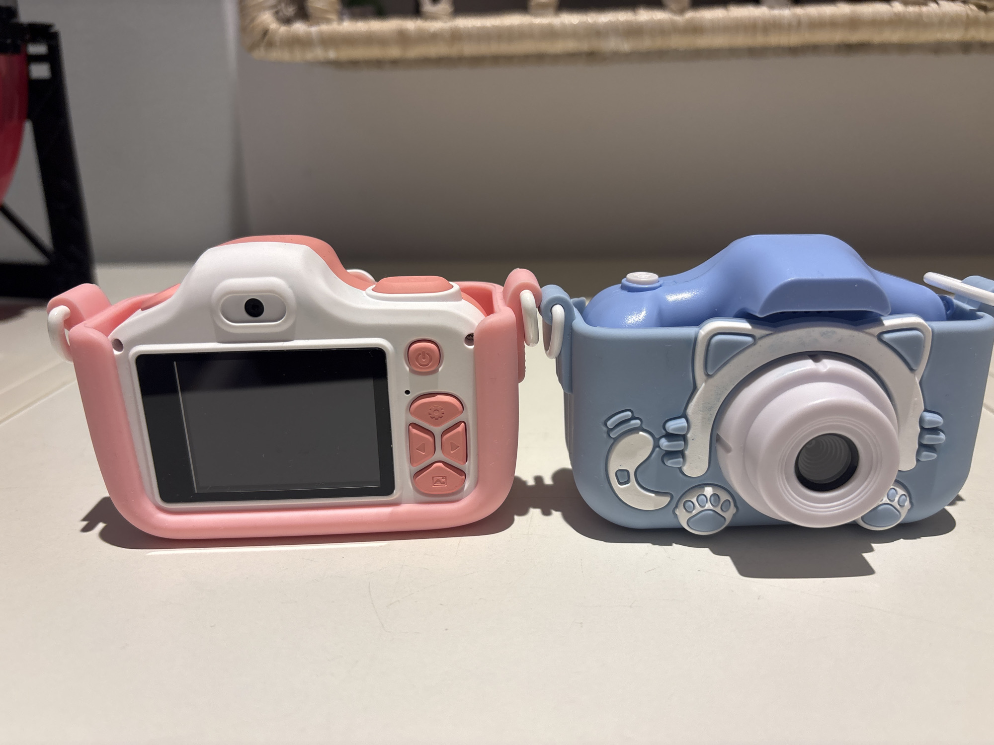 The image displays two children's digital cameras placed side-by-side on a white, reflective surface. The camera on the left is pink and white, featuring a screen, a power button, and navigation controls. The camera on the right is blue and designed to resemble a cartoon cat, with decorative ears, paws, and a central lens. The background is out of focus, showing a woven basket and a wall. The lighting suggests an indoor environment. There is no indication of the location being Dubai, United Arab Emirates, nor are there any people or actions depicted.