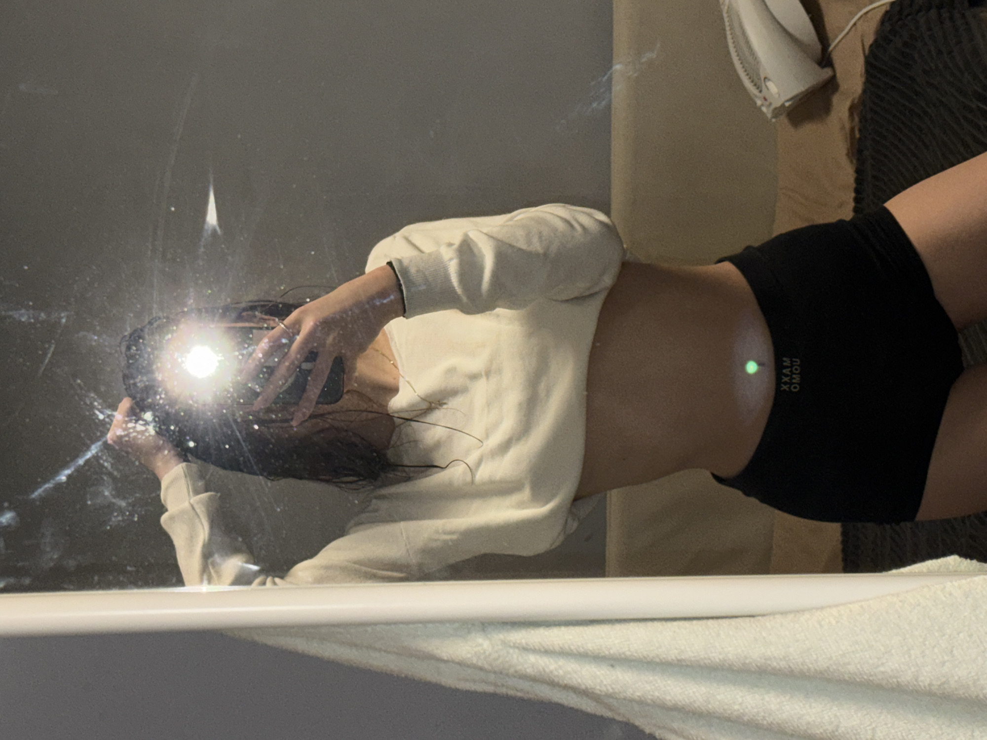 A person is reflected in a mirror in what appears to be a bathroom. The person is holding a phone, presumably taking the photo. The flash from the phone obscures part of the mirror. The person is wearing a light-colored cropped sweatshirt and black shorts. A white towel is visible at the bottom of the mirror, and a hairdryer and some other soft items are on a surface behind the person in the reflection. There is no indication of the location being Sofia, Bulgaria.