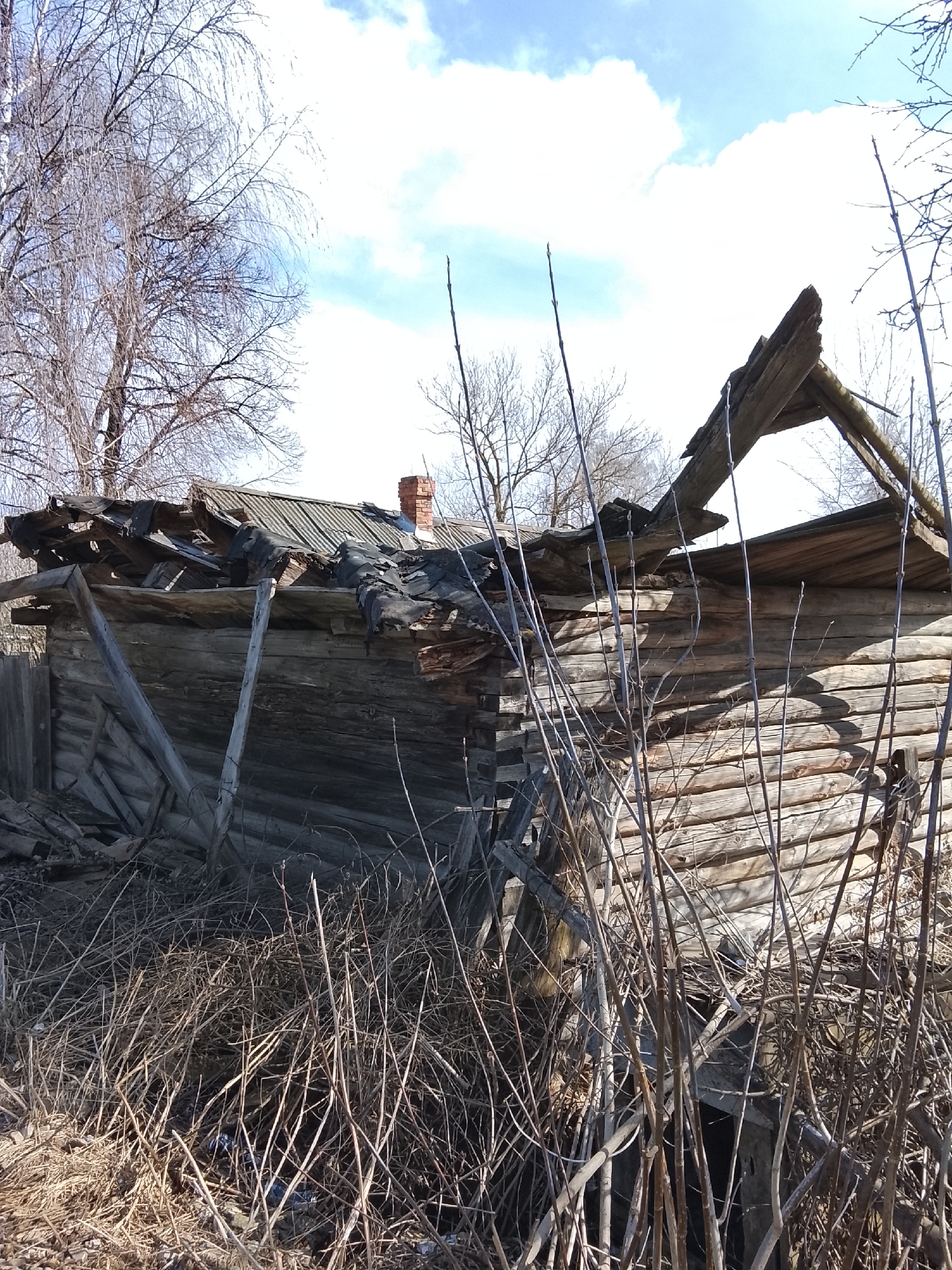 The image depicts a dilapidated wooden structure, likely a building, in an advanced state of disrepair. The roof is partially collapsed, exposing the interior. The walls are constructed of logs, weathered and worn. Debris and dead vegetation surround the structure, obscuring the base. Bare trees are visible in the background against a sky with light clouds. The presence of a chimney suggests it was once a residence, but the overall impression is one of abandonment and decay. The photograph was taken in Высокиничи, Russia.