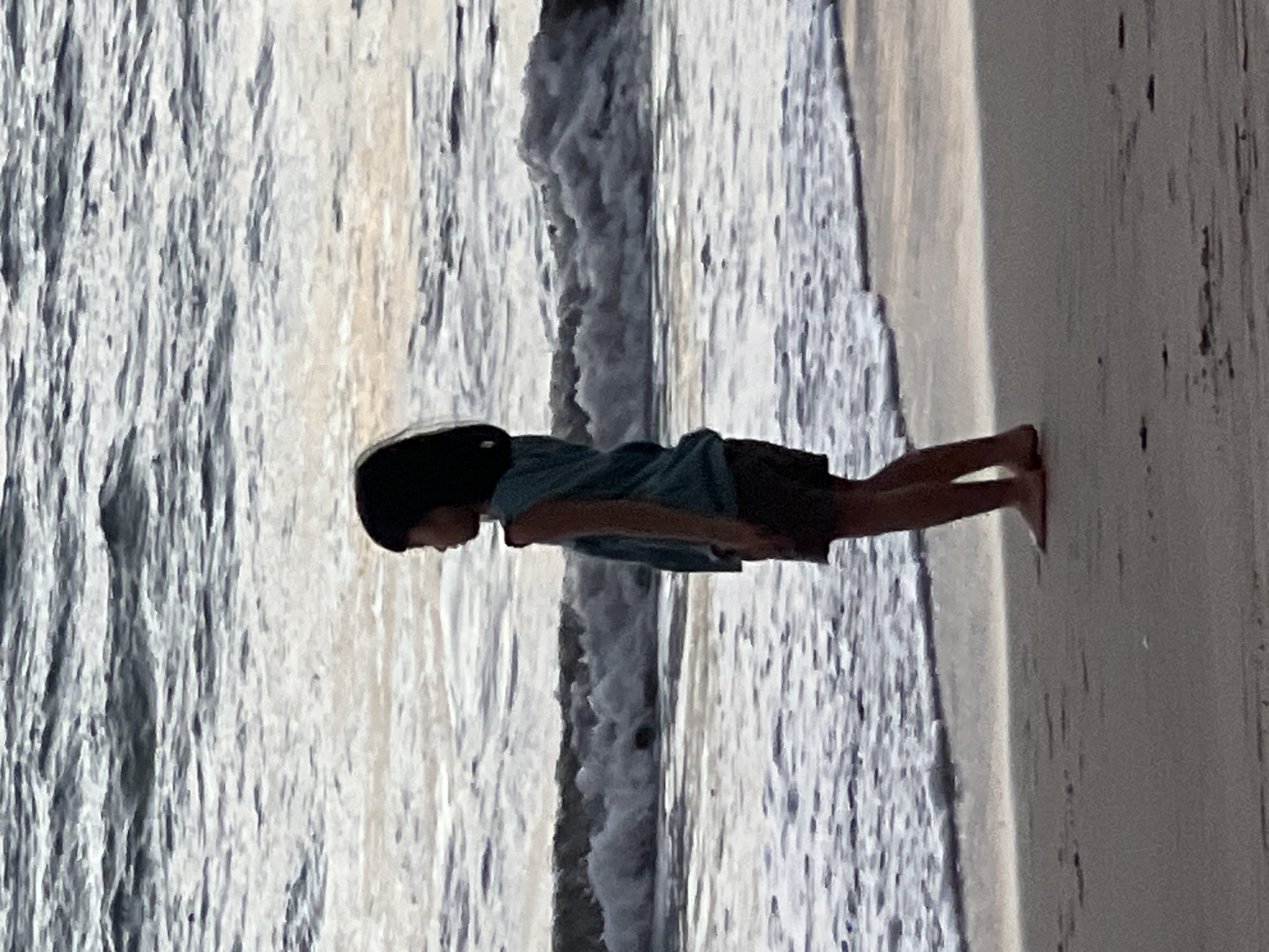A side-view medium shot captures a young child standing on the sandy shore of Kuta Beach, Indonesia, facing the ocean. The child, silhouetted against the water, wears a teal sleeveless top and dark shorts. The ocean is calm with small waves lapping at the shore, and the sky above is overcast, casting a muted light on the scene. The sand is wet near the water's edge, with footprints visible further up the beach. The overall atmosphere is serene and contemplative.