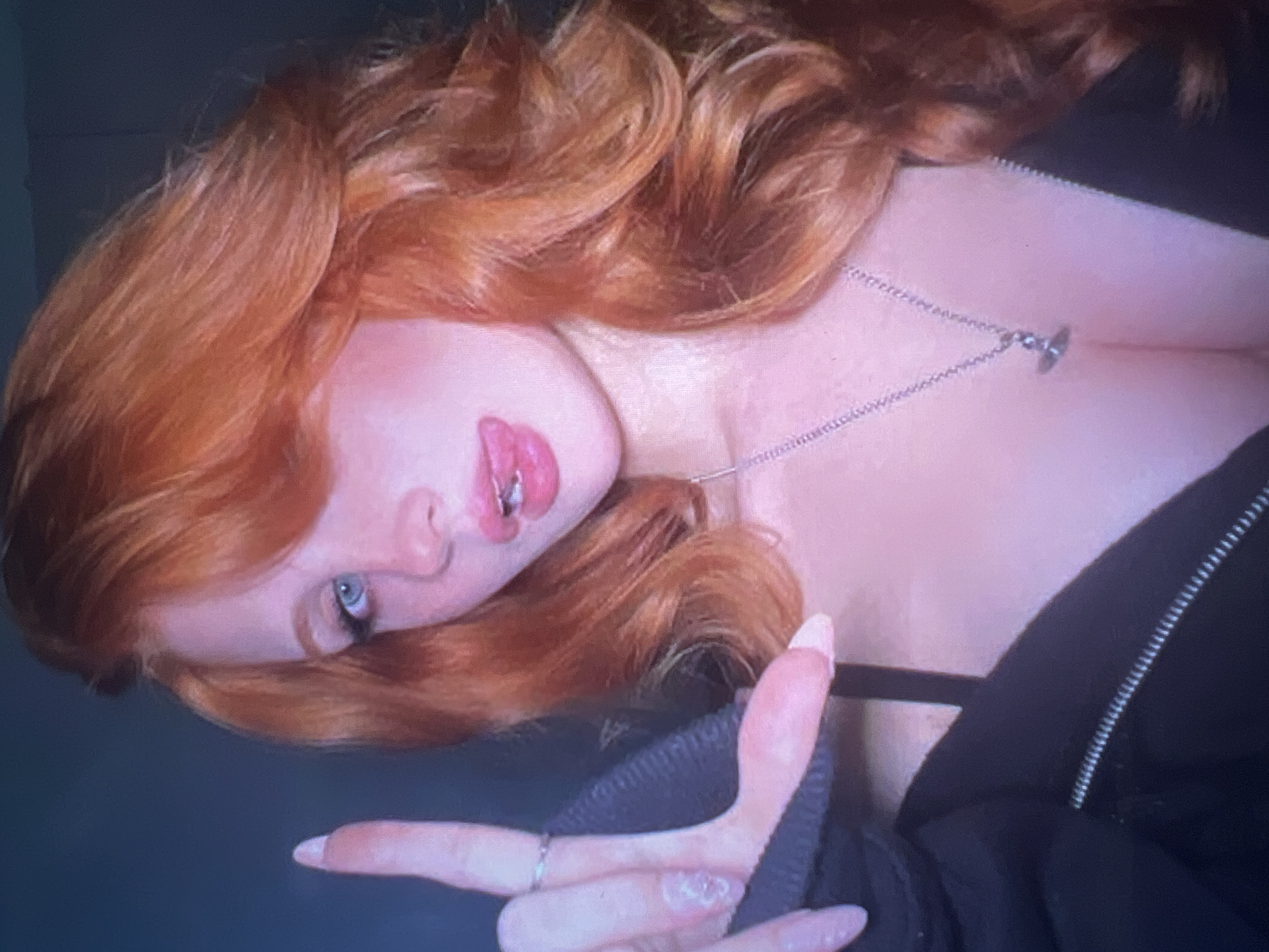 A young woman with bright red hair and blue eyes is pictured in a close-up selfie. She is wearing a black zip-up jacket and a silver necklace with a small pendant. Her tongue is sticking out slightly, and her left hand is raised, with her index finger and thumb forming an "L" shape. The background is dark and out of focus. There is no identifiable location in Minsk, Belarus.