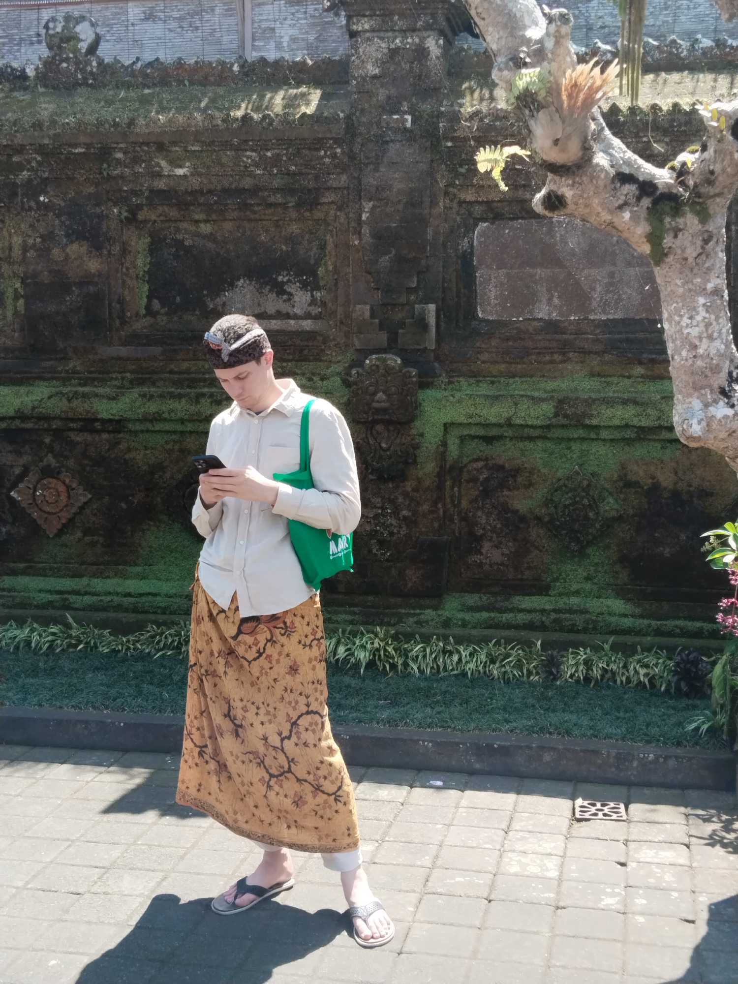 In the city of Bangli, Indonesia, a Caucasian male is standing on a paved walkway in front of a moss-covered stone wall. He is wearing a light-colored button-down shirt, a patterned sarong, a head covering, and flip-flops. He is holding a smartphone and appears to be looking at it. A bright green tote bag hangs from his left shoulder. The background features the textured stone wall, some greenery, and a tree trunk with moss and small plants growing on it. The scene is illuminated by bright sunlight, casting distinct shadows on the ground.