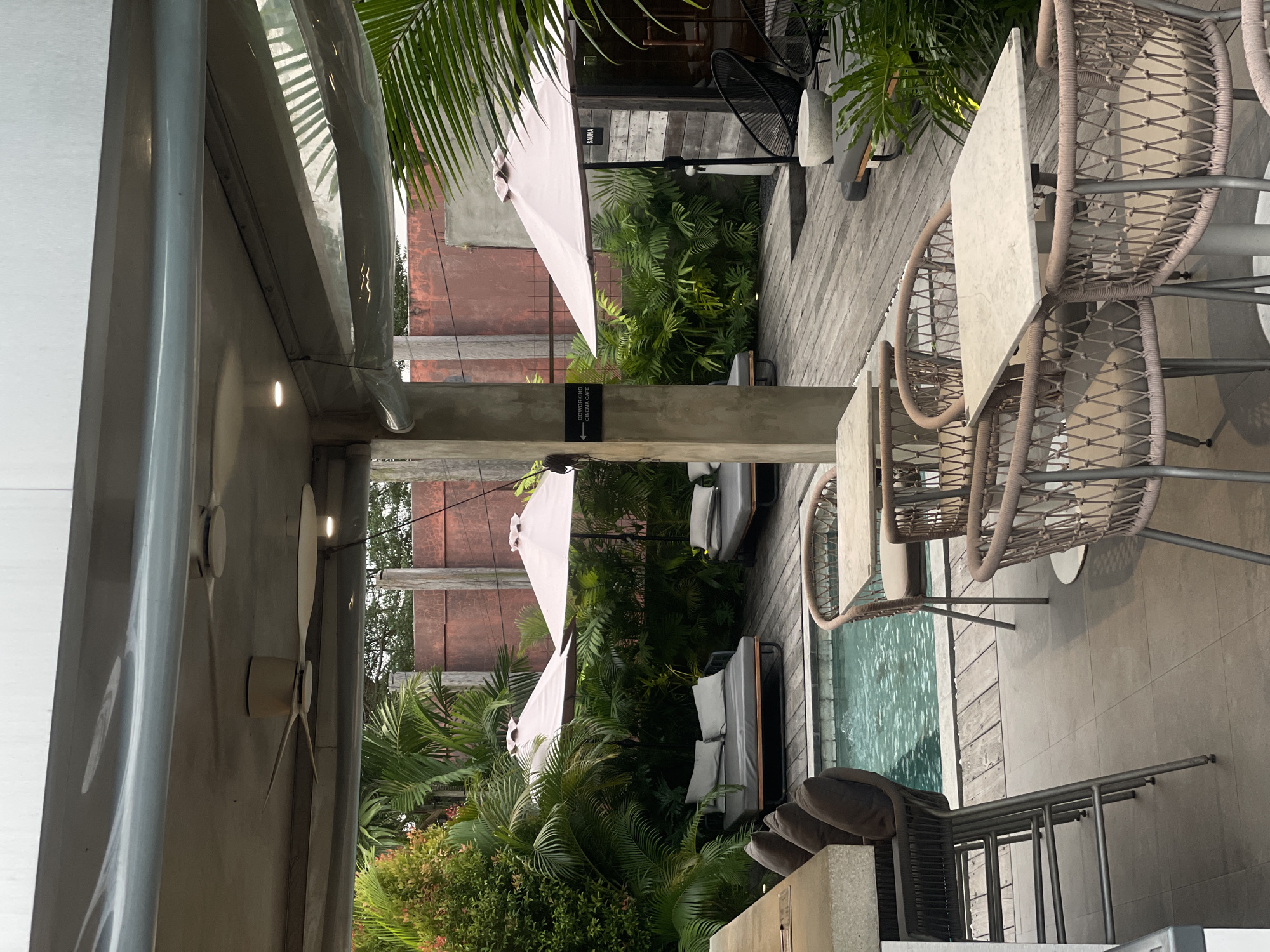 The image shows an outdoor seating area with a swimming pool and lush greenery. In the foreground, there are several tables and chairs, with some bar stools at a counter on the left. The tables are made of marble or a similar stone, and the chairs are woven with a light-colored material. A swimming pool is visible in the mid-ground, with a wooden deck surrounding it. There are several lounge chairs with cushions by the pool, and two large umbrellas providing shade. In the background, there is a building with "SAUNA" written on it, and a sign pointing to "COWORKING CINEMA CAFE". The overall atmosphere is relaxed and inviting, suggesting a resort or a high-end cafe. The location is the city of Kediri, Indonesia.