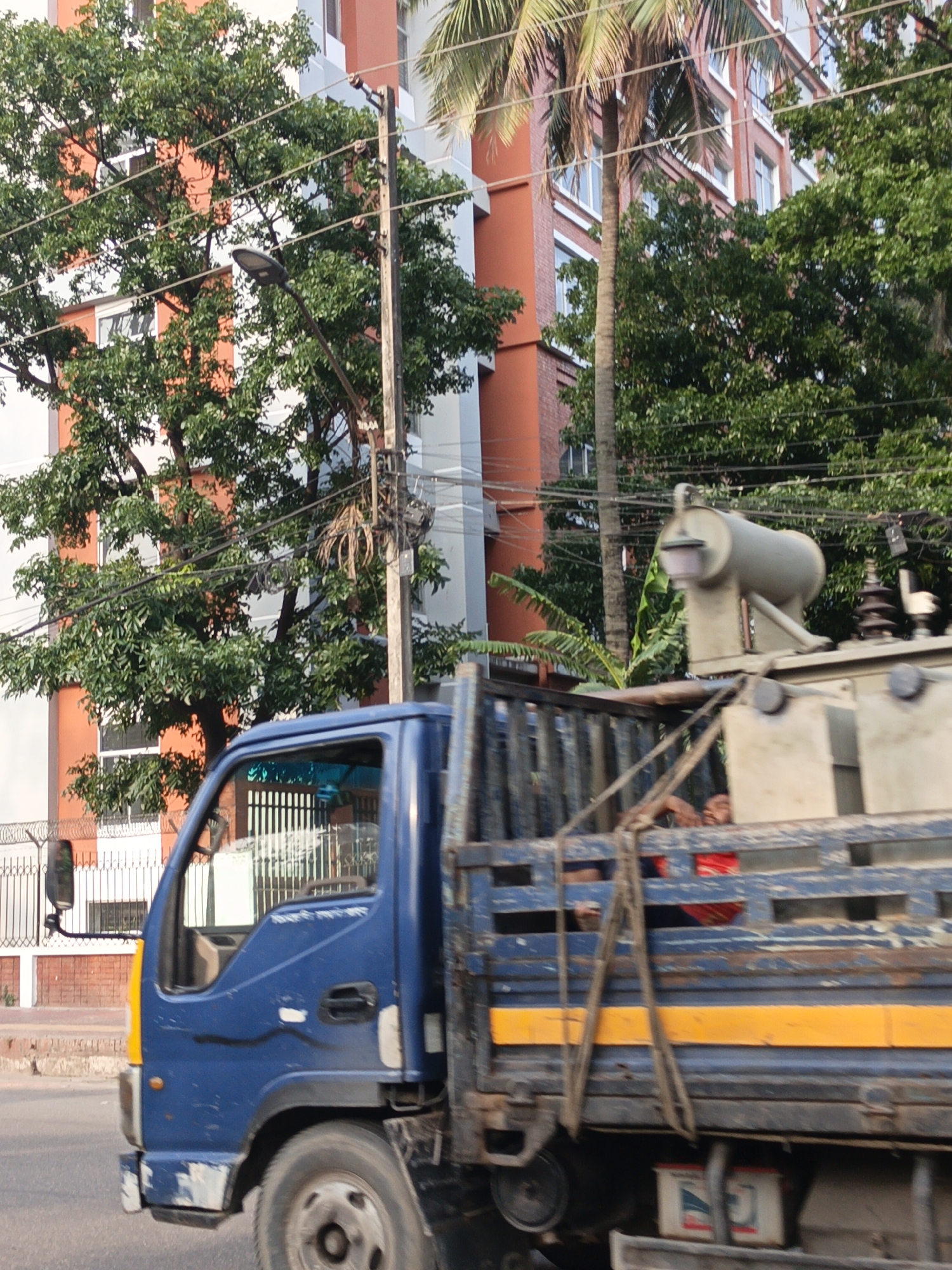 A blue flatbed truck is captured in motion on a street, viewed from a slightly elevated side-front angle. The truck's cabin is blue with a distinctive yellow stripe on its front, and minor scuffs are visible on the bumper and side. On the side of the cabin, Bengali text reads "গাড়ী নং ৬১-৯১" (Vehicle No. 61-91).
The truck's bed is carrying a large, gray industrial piece of equipment, resembling an electrical transformer or generator, secured by straps. The equipment has a prominent cylindrical component on top. Partially visible in the back of the truck, a person wearing a red shirt or garment is seen, looking towards the right side of the frame. Below the truck's bed, two white rectangular containers with red and blue logos are attached.
The background is rich with urban elements and vegetation. Multi-story buildings, painted in alternating sections of white and a warm orange-red brick color, rise behind a dense canopy of green trees. Palm trees are also visible, suggesting a warm climate. A utility pole stands prominently in the mid-ground, supporting numerous power lines and a modern-looking street light fixture.
The scene appears to be set in an urban or semi-urban environment during the daytime, under bright, natural sunlight, indicating fair weather. On the left side, a brick wall topped with a white metal fence and barbed wire is visible, suggesting a property boundary. The overall impression is of a busy street in a South Asian city, given the architecture, vegetation, and visible text.