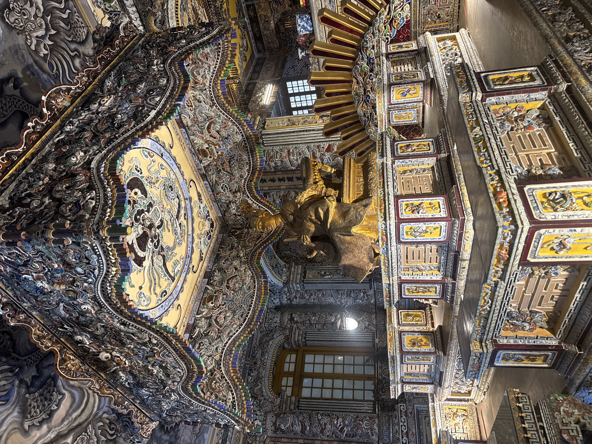 The image shows the ornate ceiling of a building, likely a temple or palace, in Thủy Xuân Ward, Vietnam. The ceiling is elaborately decorated with intricate carvings, paintings, and mosaics. The artwork depicts various figures and scenes, possibly from Buddhist or Taoist mythology. A golden statue is visible, suggesting the religious significance of the building. The architectural style is traditional Vietnamese, with a blend of Chinese and local influences. Windows provide natural light, illuminating the detailed artwork.