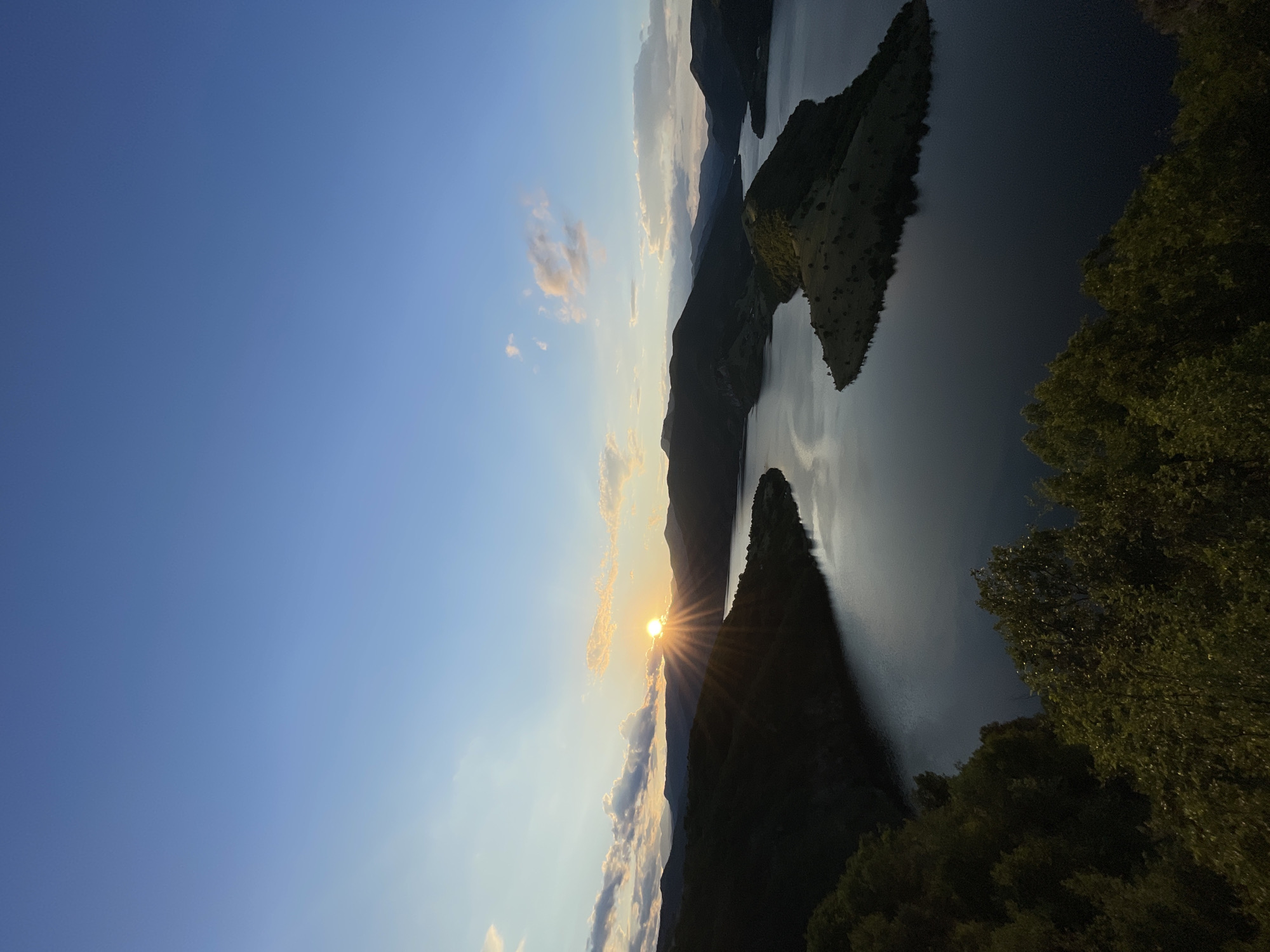 This image captures a serene sunset over a large lake surrounded by lush, dark green hills. The sun, a bright orange orb, is just beginning to dip below the horizon, casting long rays of light across the water and sky. Wispy clouds are scattered across the twilight sky, tinged with pink and orange from the setting sun. The lake itself is calm and reflects the colors of the sky and the surrounding landscape. The foreground is dominated by the dense foliage of trees, which are silhouetted against the dramatic sky and water. The location is the dam on the Borovitsa River, near the town of Borovitsa in Bulgaria. There are no people or specific objects of note visible in the scene.