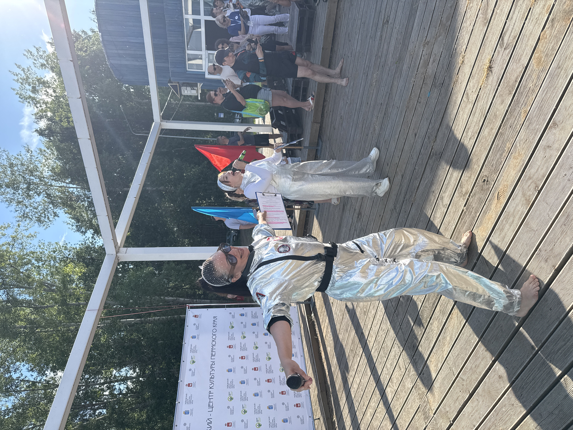 The image shows a group of people gathered outdoors on a wooden deck. In the foreground, a woman dressed in a shiny silver astronaut costume, complete with a helmet and sunglasses, is holding a microphone and appears to be speaking or performing. She is barefoot. Behind her, another woman in a white top and silver pants stands holding a clipboard. A red flag is visible to the left of the performers, and a blue flag is partially visible at the bottom left. In the background, other people are standing and watching, some holding phones. A building with blue siding is visible behind the crowd, and trees and a bright blue sky are in the background, suggesting a sunny day. The scene appears to be an outdoor event or festival. Visible text on a banner at the bottom of the image reads "ЦЕНТР КУЛЬТУРЫ ПЕРМСКОГО КРАЯ" which translates to "CENTER OF CULTURE OF PERM KRAI", and indicates the location is likely Chaikovsky, Russia. The overall mood seems festive and engaging.