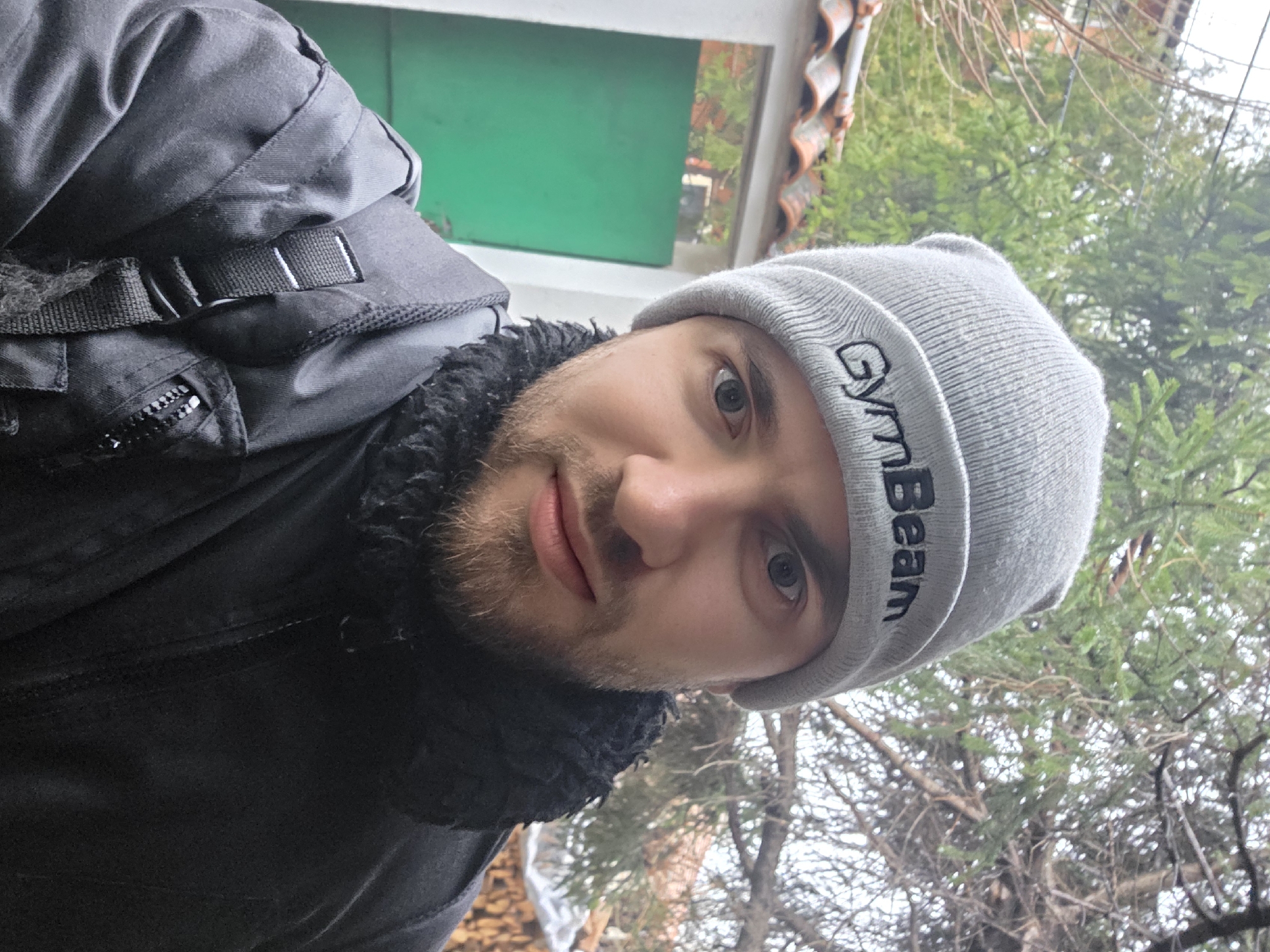 A man with a beard and blue eyes, wearing a grey "GymBeam" beanie and a black winter jacket, is taking a selfie. He is outdoors, and there are bare tree branches and green foliage visible in the background. There is a building element with green and brown coloration as well, possible a window frame and roof tile, visible behind and above him. I have no means to provide the location in the city of Yagodovo, Bulgaria.