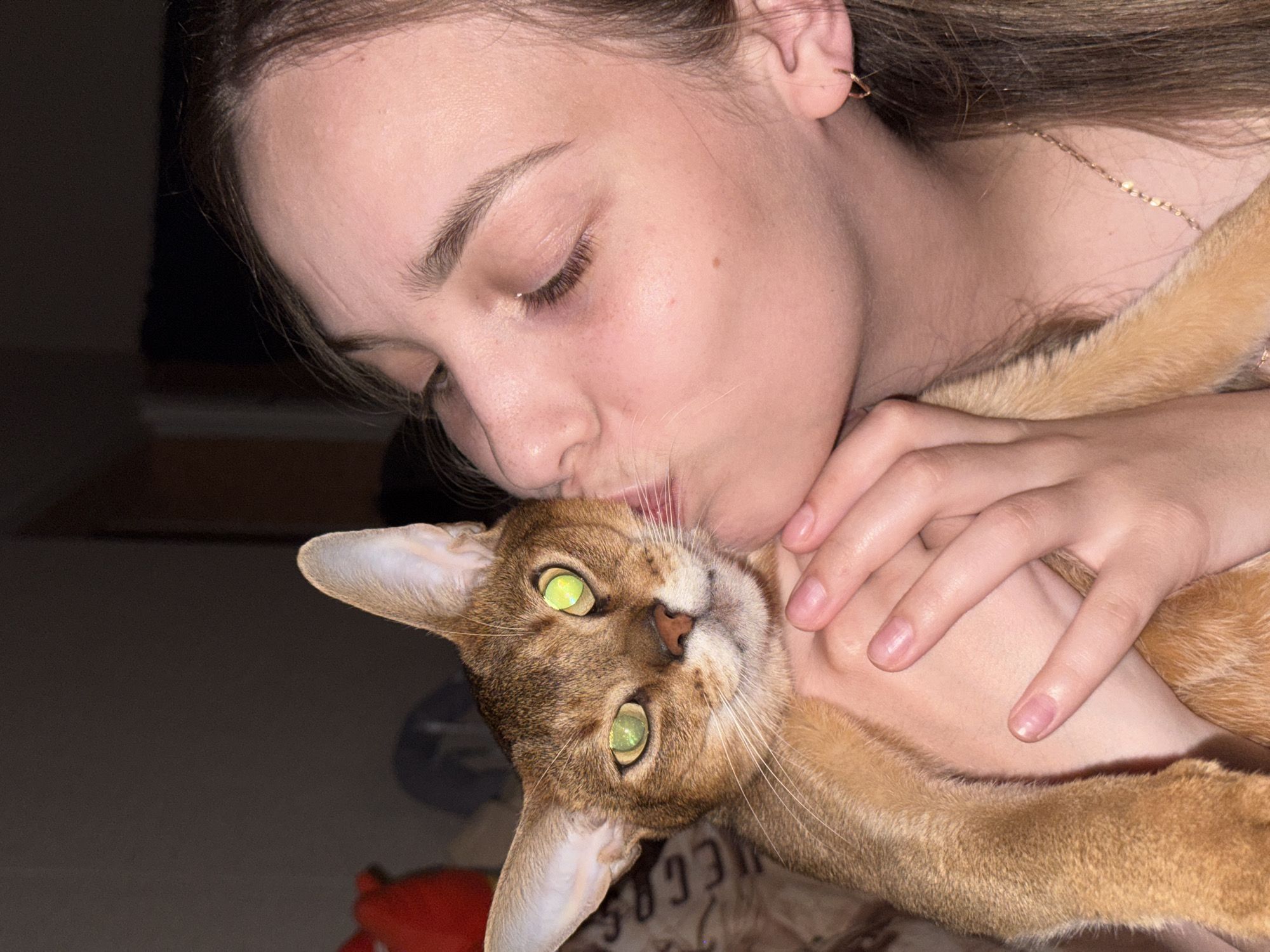 A young woman with brown hair is holding an Abyssinian cat close to her face, kissing it on the cheek. The cat has striking green eyes and a golden-brown coat. The background is dark and out of focus, with hints of objects that suggest an indoor setting. The woman is wearing a necklace and earrings. The image appears to be a personal photograph, possibly taken in a domestic environment in Chaikovsky, Russia.