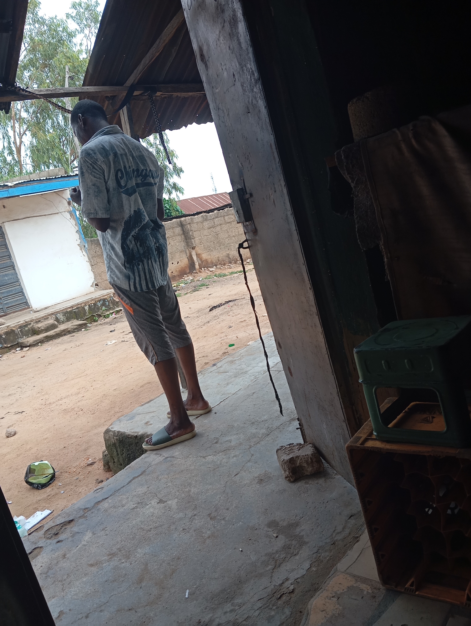 In Jalingo, Nigeria, a person stands at the entrance of a building. They are wearing a grey t-shirt with a graphic print and grey shorts, and are barefoot. The building appears to be constructed with corrugated metal roofing and brick walls. The ground is sandy and uneven. To the right of the entrance, there is a stack of plastic crates, one of which is green and the other is brown and appears to be empty. In the foreground, there is a concrete slab and a green object, possibly a cap, on the ground. The overall scene depicts an outdoor urban environment.