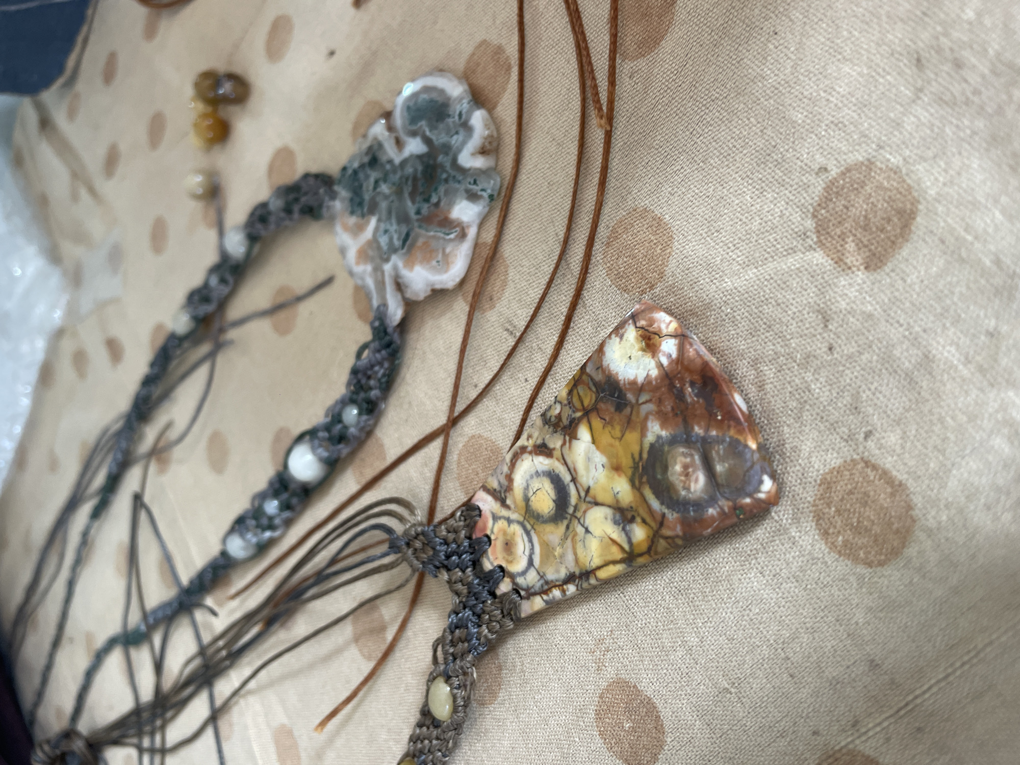 The image shows two macrame necklaces with gemstone pendants. The necklaces are laid out on a light-colored fabric with a pattern of darker brown polka dots. The pendant on the left is a flat, elongated stone with a complex pattern of yellow, brown, and white. The pendant on the right is a larger, more irregular shaped stone with white, gray, and green marbled patterns. Both necklaces have macrame cords and some small, light-colored beads. Several thin, brown cords are also visible, lying across the fabric and extending from the necklaces. The lighting suggests an indoor setting, likely a craft or workspace. There are no people visible in the image. The location of Bhagsu, India is not depicted or suggested by the image.