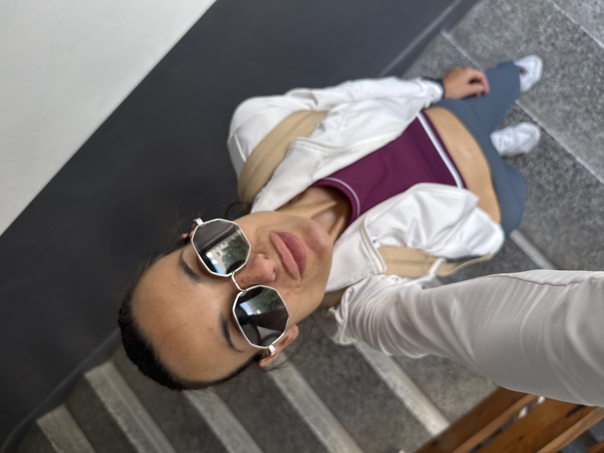 An individual is taking a selfie on a concrete staircase with gray steps and dark gray walls. The person is wearing a white jacket over a purple sports bra, gray pants, and white sneakers. They are also wearing large, angular sunglasses. A tan backpack is visible over their shoulders. The scene appears to be outdoors, possibly in an urban setting like Pleven, Bulgaria.