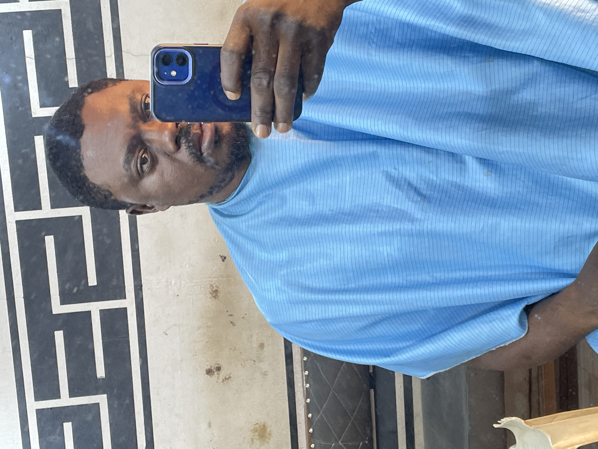 A man with dark skin, a dark beard, and short dark hair takes a selfie in what appears to be a barber's shop or salon. He holds a dark blue smartphone in his right hand. He is wearing a light blue barber's cape. The reflection in the background suggests he is in a room with decorative wall panels, likely the interior of the shop. It is not possible to confirm the location is in Jos, Nigeria, from the image alone.