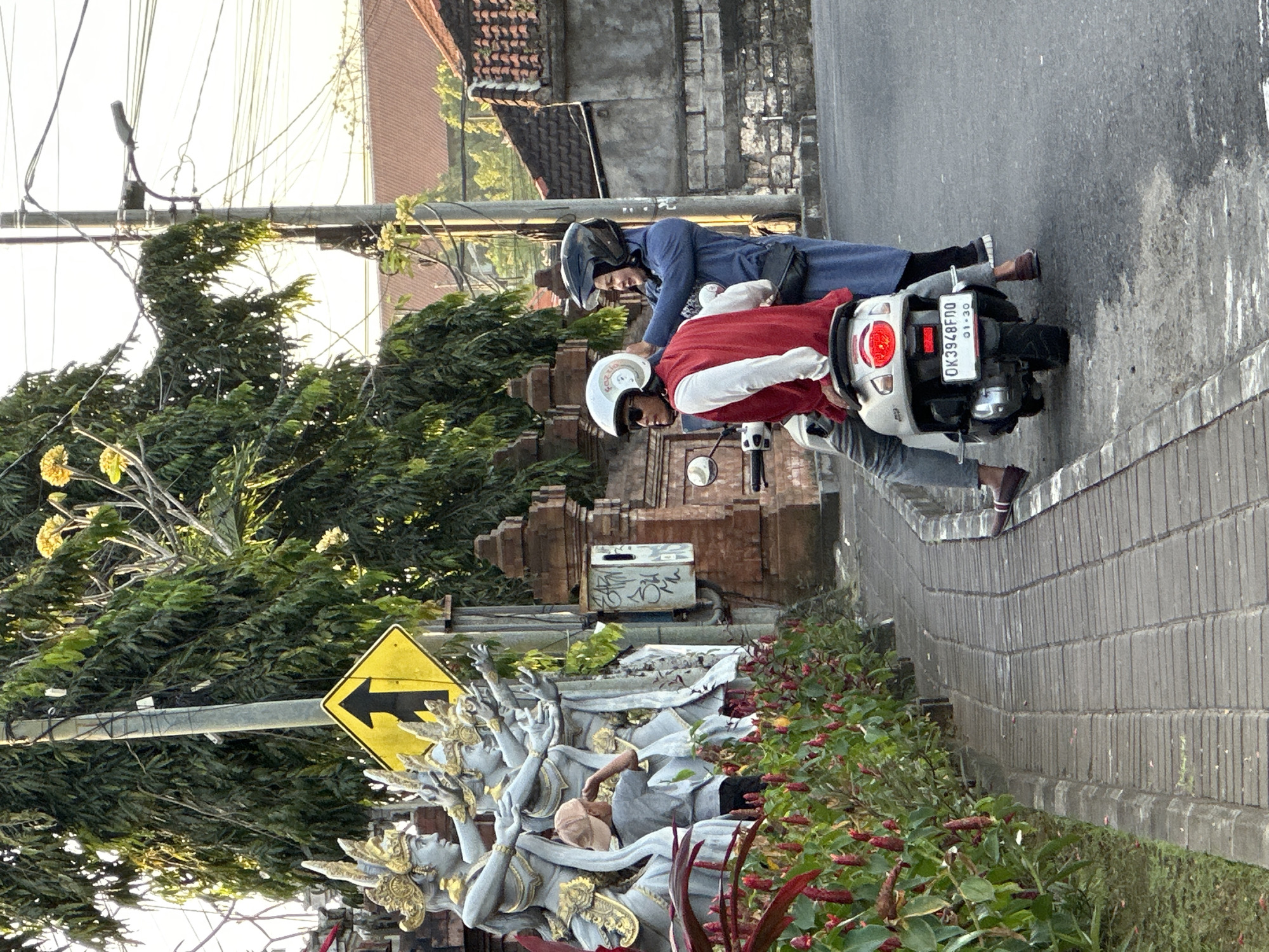 Two people on a scooter are stopped on a street, possibly waiting or taking a break. The person in the back, wearing a blue top and helmet, is leaning forward, seemingly talking to the driver. The driver, wearing a red and white jacket and a white helmet, is looking forward. The scooter has a license plate that reads "OK3948FDO" with "01-30" below it.
The scene appears to be in a tropical or subtropical location, indicated by the lush green foliage and vibrant red flowers bordering the path. A yellow diamond-shaped sign with a black arrow pointing left is visible on a pole, suggesting a turn or specific direction. Behind the scooter, a brick wall and building with a tiled roof are present. To the left, a large, ornate sculpture with multiple figures and golden accents is partially visible, hinting at a cultural or decorative element in the vicinity.
The lighting suggests it is either early morning or late afternoon, with soft sunlight casting shadows. The overall atmosphere is relaxed and casual, with no apparent urgency or distress. The location cues, such as the architecture and foliage, combined with the Indonesian license plate, point to Kuta Selatan, Indonesia.