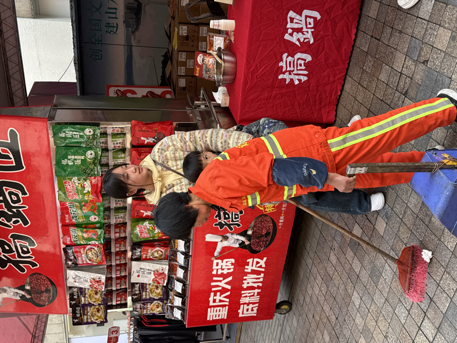 A street vendor in an orange jumpsuit sweeps the sidewalk next to a red food cart advertising "Chongqing Hot Pot Base Material Wholesale." The cart is stocked with packaged hot pot ingredients. A woman and child stand nearby, seemingly customers. The cart has signage in Chinese. Above the cart is a red table with more merchandise and a banner that reads "Gao Guo Jiang." The scene is a bustling outdoor market.
