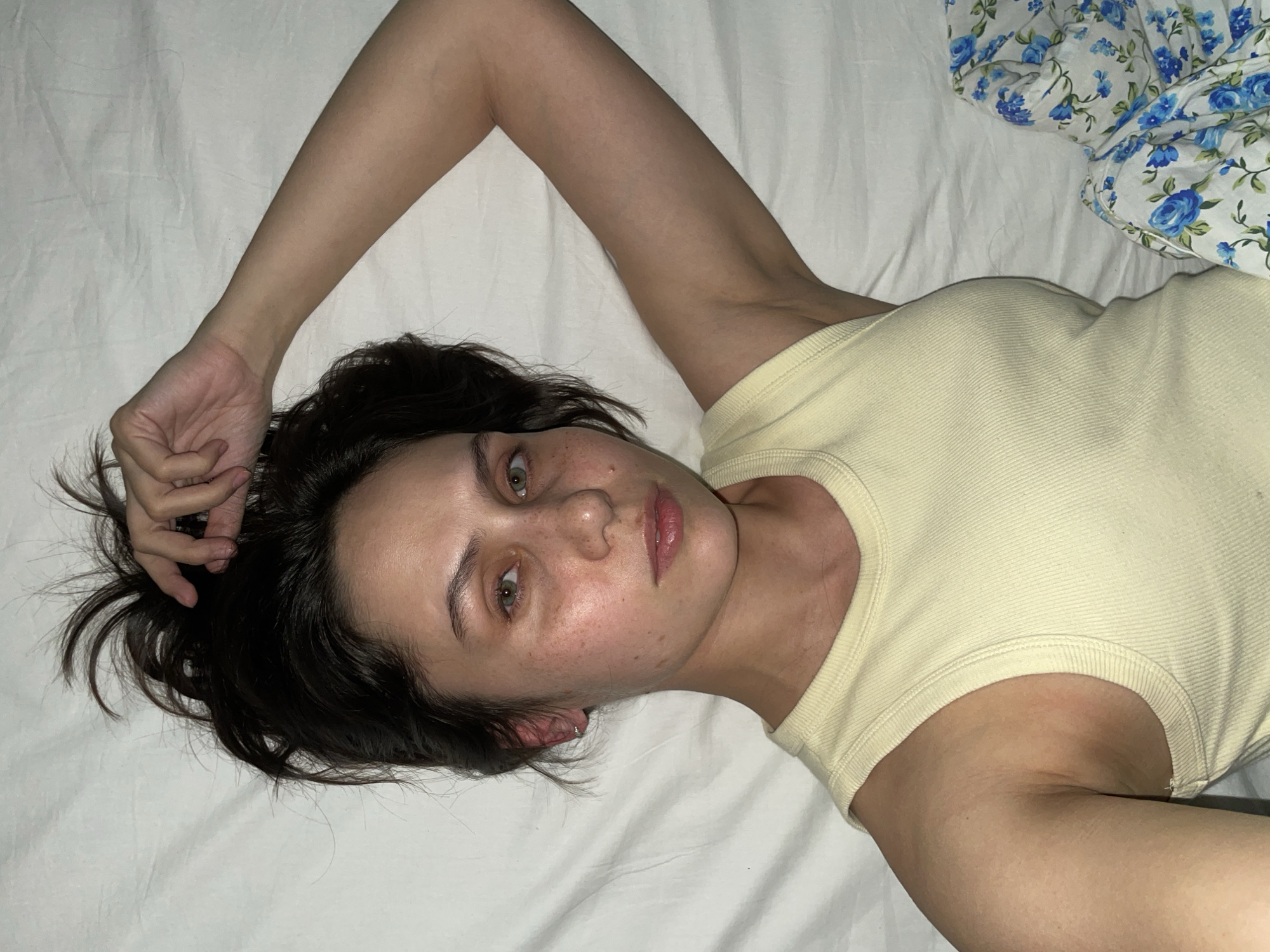 A young woman is depicted lying on her back, looking directly at the viewer. She has dark, medium-length hair fanned out around her head against a white sheet. Her right arm is raised and bent, with her hand resting near her head and fingers gently touching her hair, while her left arm is positioned alongside her body. She is wearing a light yellow ribbed tank top.
Her complexion is fair, with noticeable freckles scattered across her nose and cheeks. Her eyes are a striking light green or hazel, and her lips are slightly parted with a natural, soft appearance. Her expression appears contemplative, possibly tired or reflective, maintaining direct eye contact with the camera.
The background consists primarily of white, rumpled bedding, likely a bed sheet. In the upper right corner of the frame, a portion of a crumpled white blanket or duvet with a blue floral pattern, featuring roses and leaves, is visible. The lighting is soft and indirect, suggesting an indoor environment, possibly in the evening or early morning, casting gentle shadows and highlights on her face and body. There is no visible text in the image. This intimate and relaxed scene is set in Izhevsk, Russia.